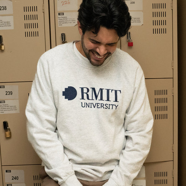 Model positioned in front of lockers wearing a white marle RMIT Essential Jumper, adorned with a navy RMIT University logo. The backdrop of lockers provides a collegiate atmosphere, emphasising the sweater's casual appeal. Available at the RMIT Store, perfect for students and alumni.