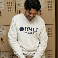 Model positioned in front of lockers wearing a white marle RMIT Essential Jumper, adorned with a navy RMIT University logo. The backdrop of lockers provides a collegiate atmosphere, emphasising the sweater's casual appeal. Available at the RMIT Store, perfect for students and alumni.