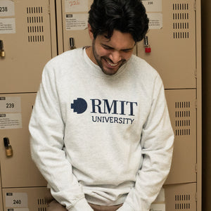 Model positioned in front of lockers wearing a white marle RMIT Essential Jumper, adorned with a navy RMIT University logo. The backdrop of lockers provides a collegiate atmosphere, emphasising the sweater's casual appeal. Available at the RMIT Store, perfect for students and alumni.