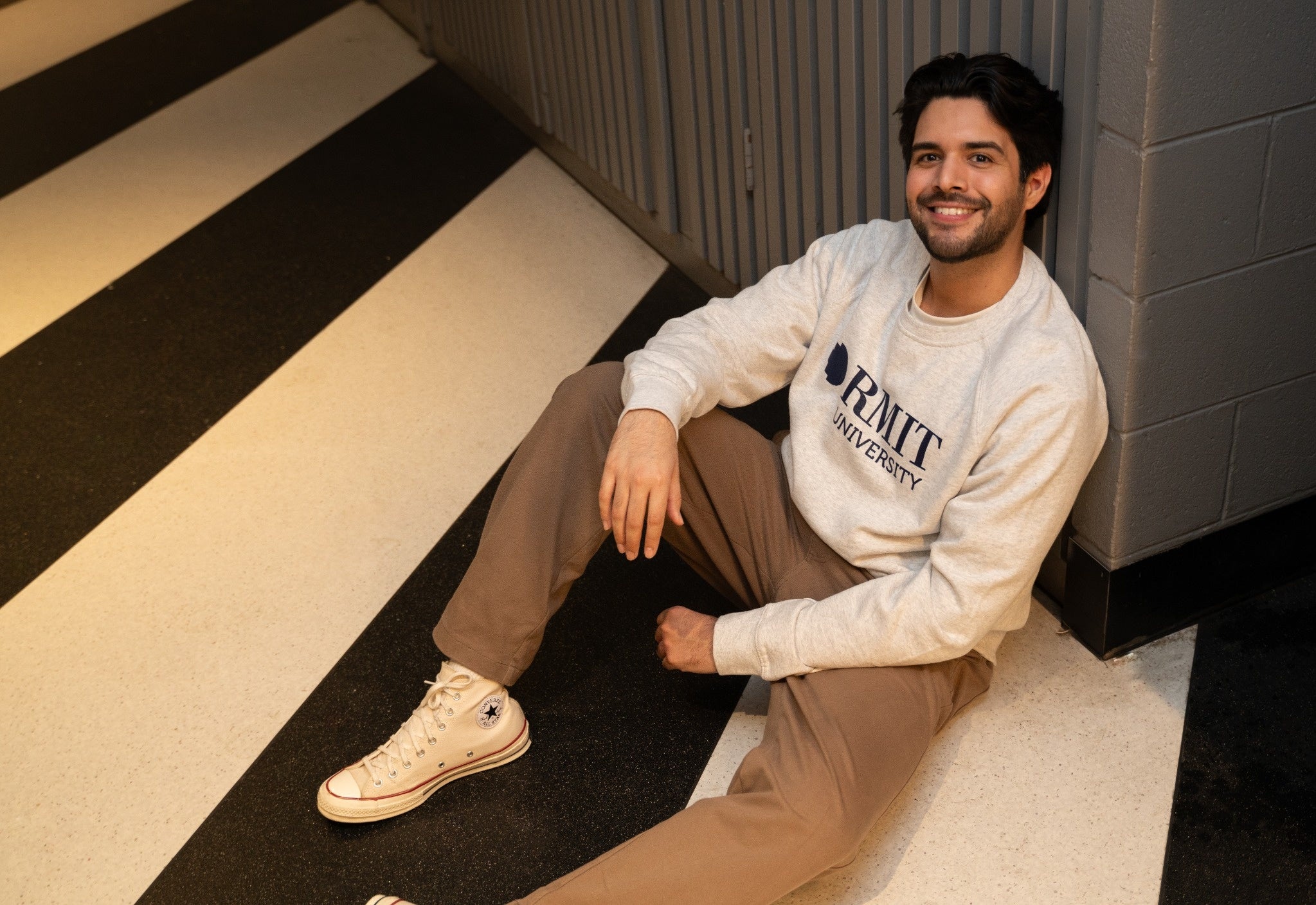 Essential Jumper in white marle, full body lifestyle view worn by a model seated on an indoor staircase with black and cream striped steps, featuring a soft fleece fabric with subtle grey marle flecks, ribbed cuffs and hem, crew neckline, and a bold navy RMIT University logo printed at the centre chest, styled with beige trousers and cream high-top sneakers in a warm indoor setting, available at the RMIT Store.