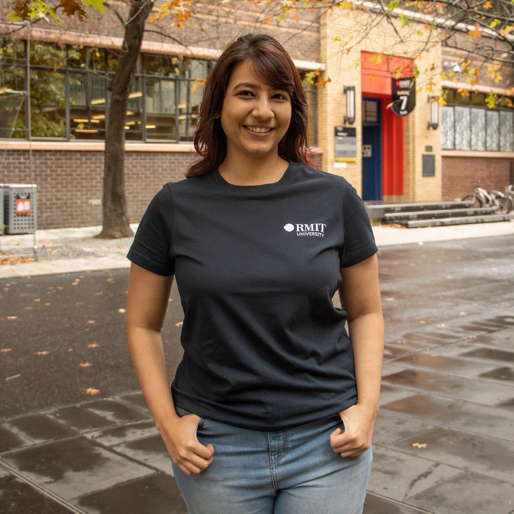 Model wears RMIT Essential Tee in navy, displaying the RMIT University logo, paired with jeans for an outdoor setting. Ideal for showcasing school pride; available at the RMIT Store.