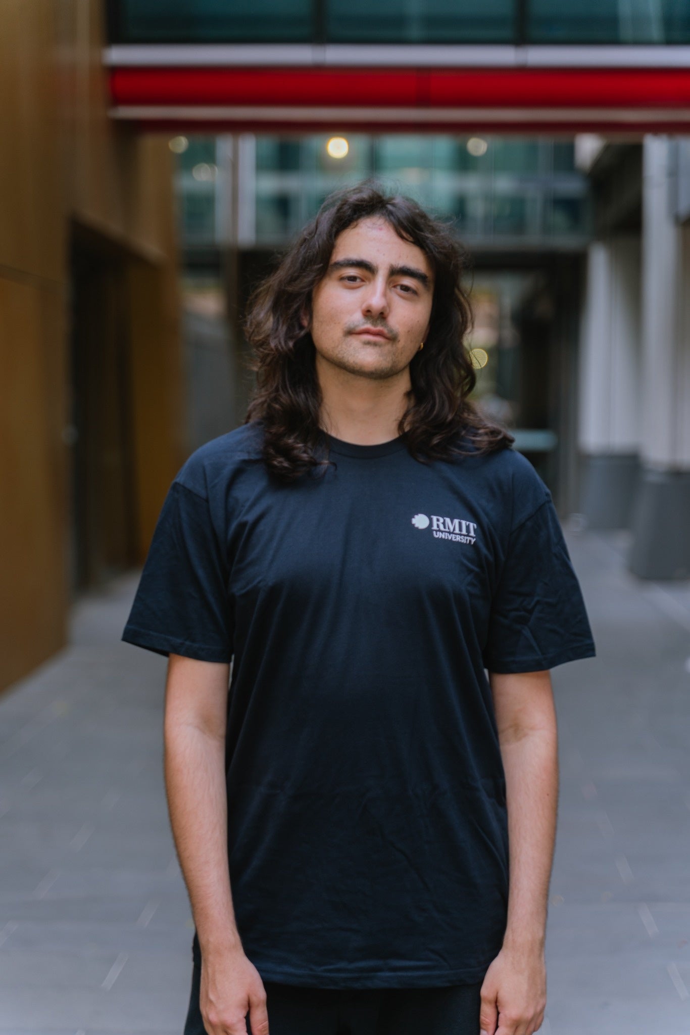 Essential Tee in navy blue, front view, worn by a model standing facing the camera in an outdoor laneway with soft-focus buildings and a red overhead beam, featuring a small white RMIT University logo printed on the left chest, short sleeves, crew neckline, and relaxed cotton fit, available at the RMIT Store.