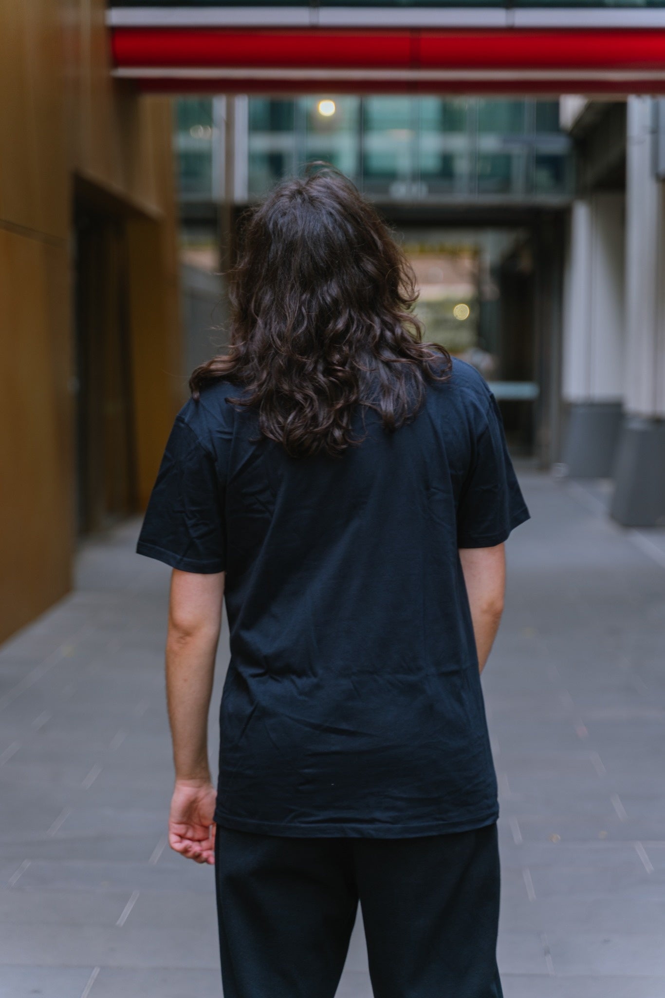 Essential Tee in navy blue, rear view, worn by a model standing facing away in an outdoor urban laneway setting with blurred architectural background and a red overhead beam, featuring a plain back with no visible branding, short sleeves, crew neckline, and soft cotton fabric drape, available at the RMIT Store.