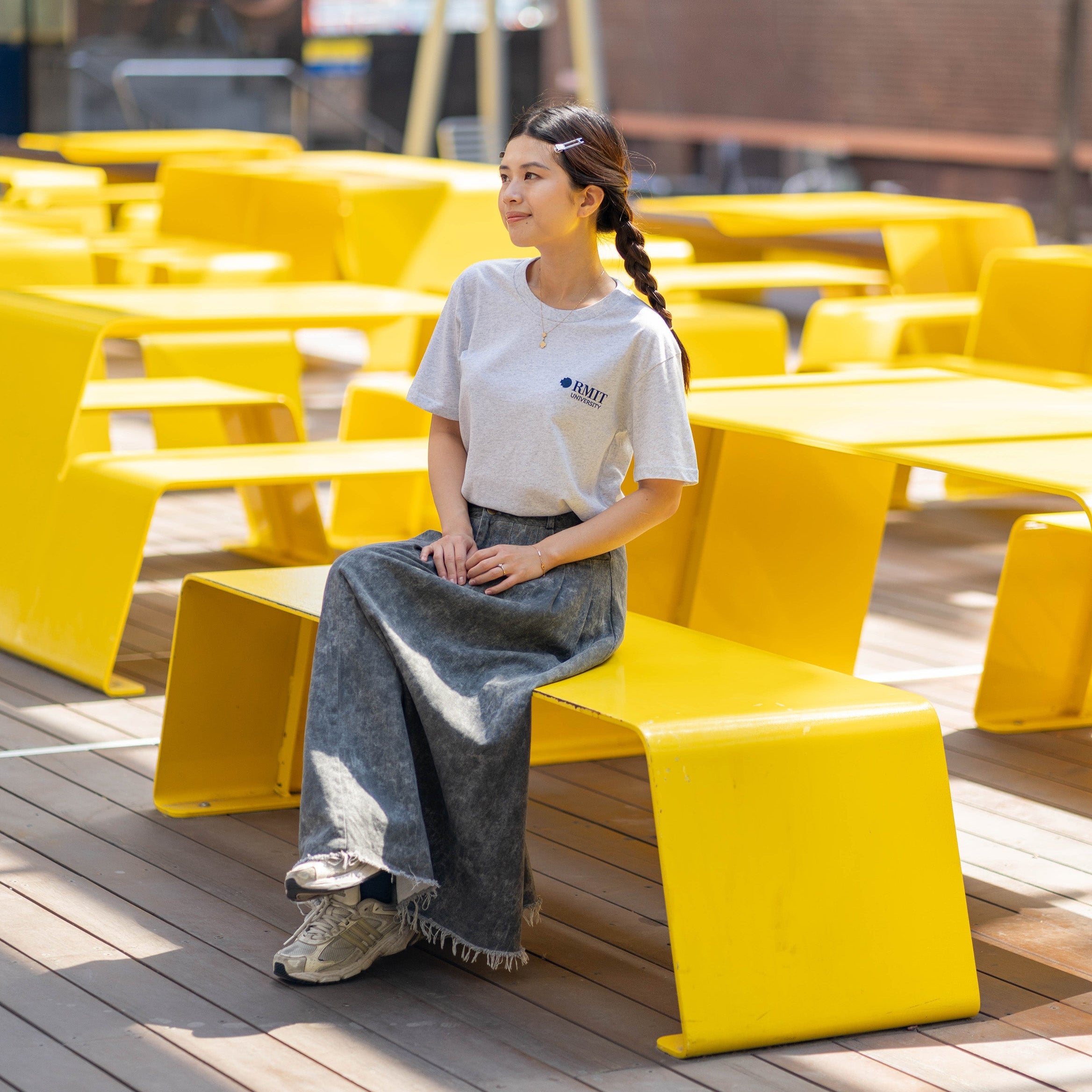 Model wears RMIT Essential Tee in white marle with RMIT University branding on the chest, paired with a long denim skirt while seated on a bright yellow bench. Available at the RMIT Store.