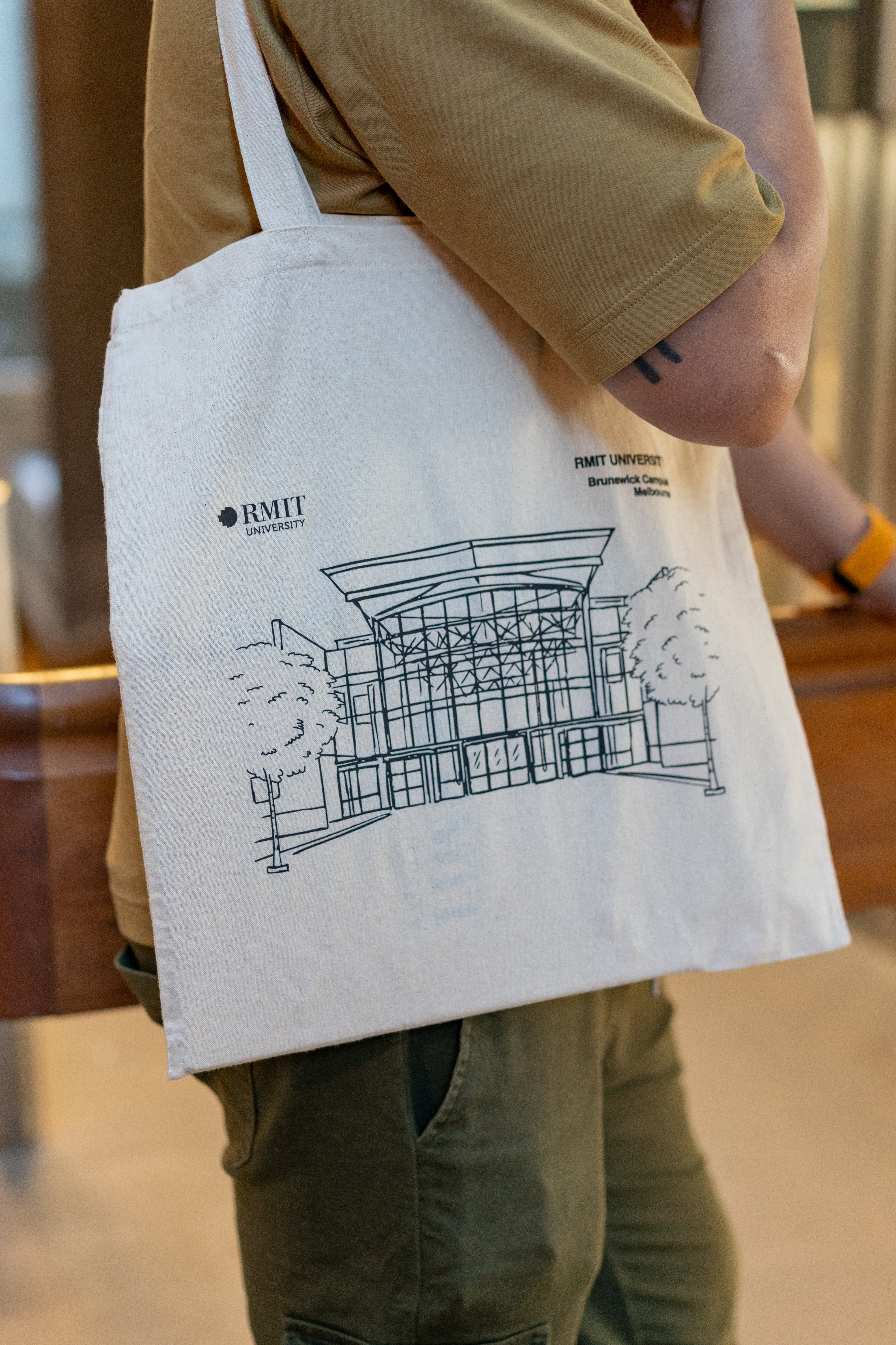 Close-up side view of a model wearing the RMIT Store Everyday Campus Tote in the Brunswick Campus variant. The natural cotton tote features a detailed black line art illustration of the Brunswick Campus building with its prominent glass entry and symmetrical design, flanked by trees. The RMIT University logo and campus name are printed above the artwork. The model wears a light brown T-shirt and olive trousers, shown indoors with soft background lighting.