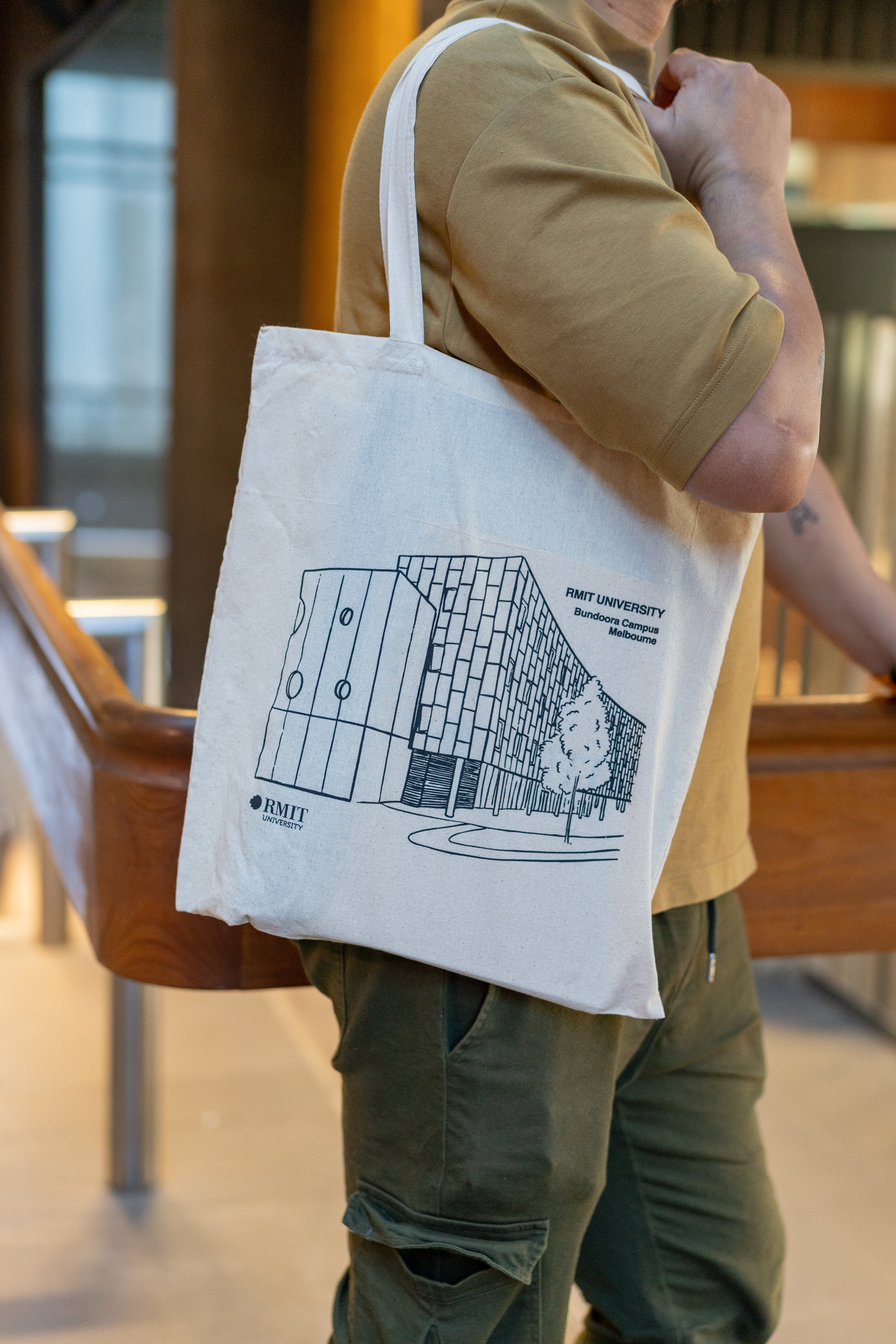 Side view of a model carrying the RMIT Store Everyday Campus Tote in the Bundoora Campus variant. The natural cotton bag showcases a bold black line art illustration of the Bundoora Campus building with a distinctive angled façade, circular window accents, and adjacent trees. The RMIT University logo and campus name appear in black print. The model wears a tan polo shirt and olive green cargo pants, photographed indoors with warm lighting.