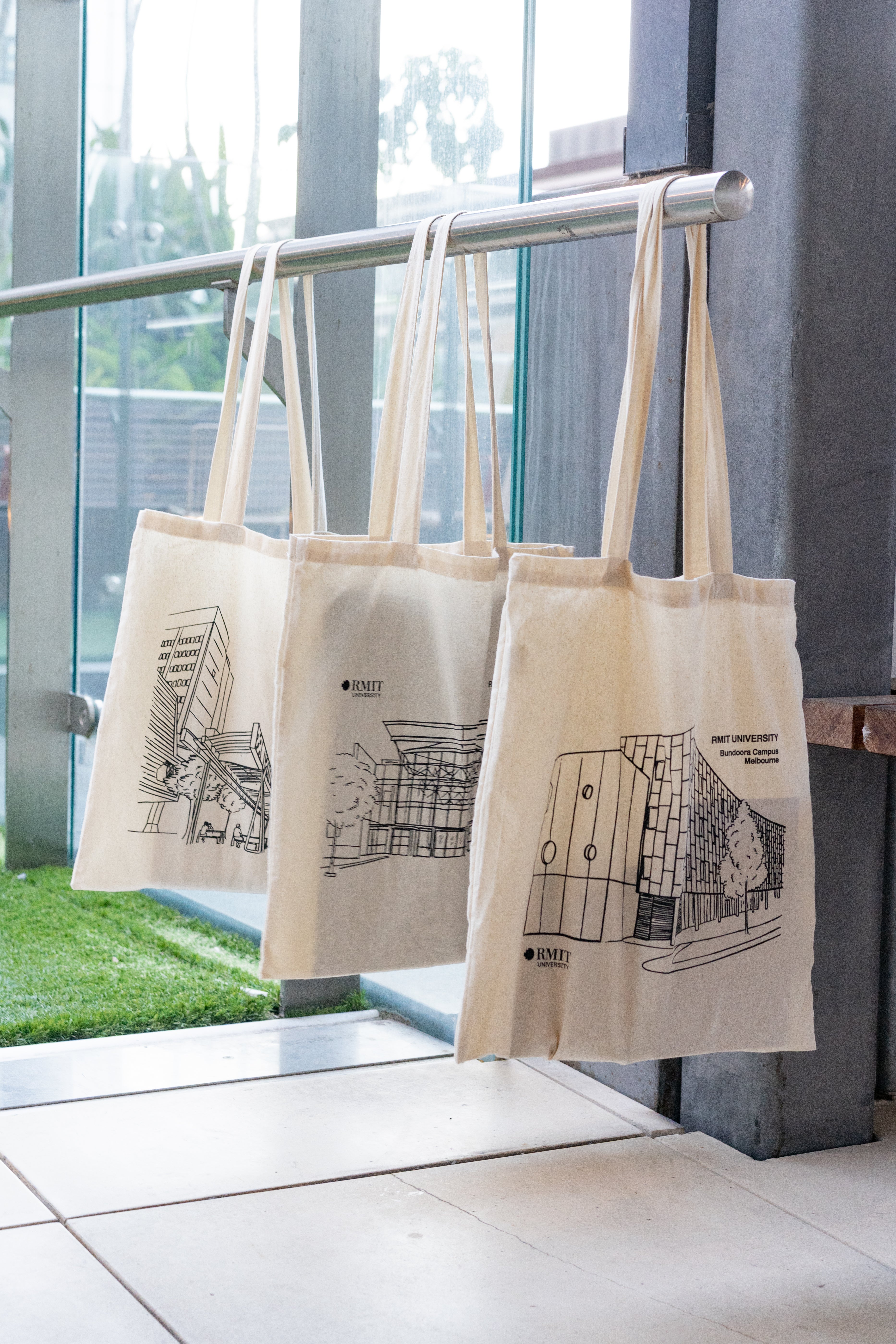 Three natural cotton RMIT Store Everyday Campus Totes hanging from a silver rail, each featuring a black line art illustration of a different RMIT University campus building. From left to right: the City Campus with trees and detailed foreground structure, Brunswick Campus with its large glass façade and surrounding trees, and Bundoora Campus with a geometric façade and circular window designs. Each tote displays the RMIT University logo and campus name in black print, set indoors by a glass wall and art.