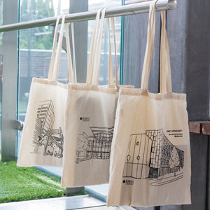Three natural cotton RMIT Store Everyday Campus Totes hanging from a silver rail, each featuring a black line art illustration of a different RMIT University campus building. From left to right: the City Campus with trees and detailed foreground structure, Brunswick Campus with its large glass façade and surrounding trees, and Bundoora Campus with a geometric façade and circular window designs. Each tote displays the RMIT University logo and campus name in black print, set indoors by a glass wall and art.