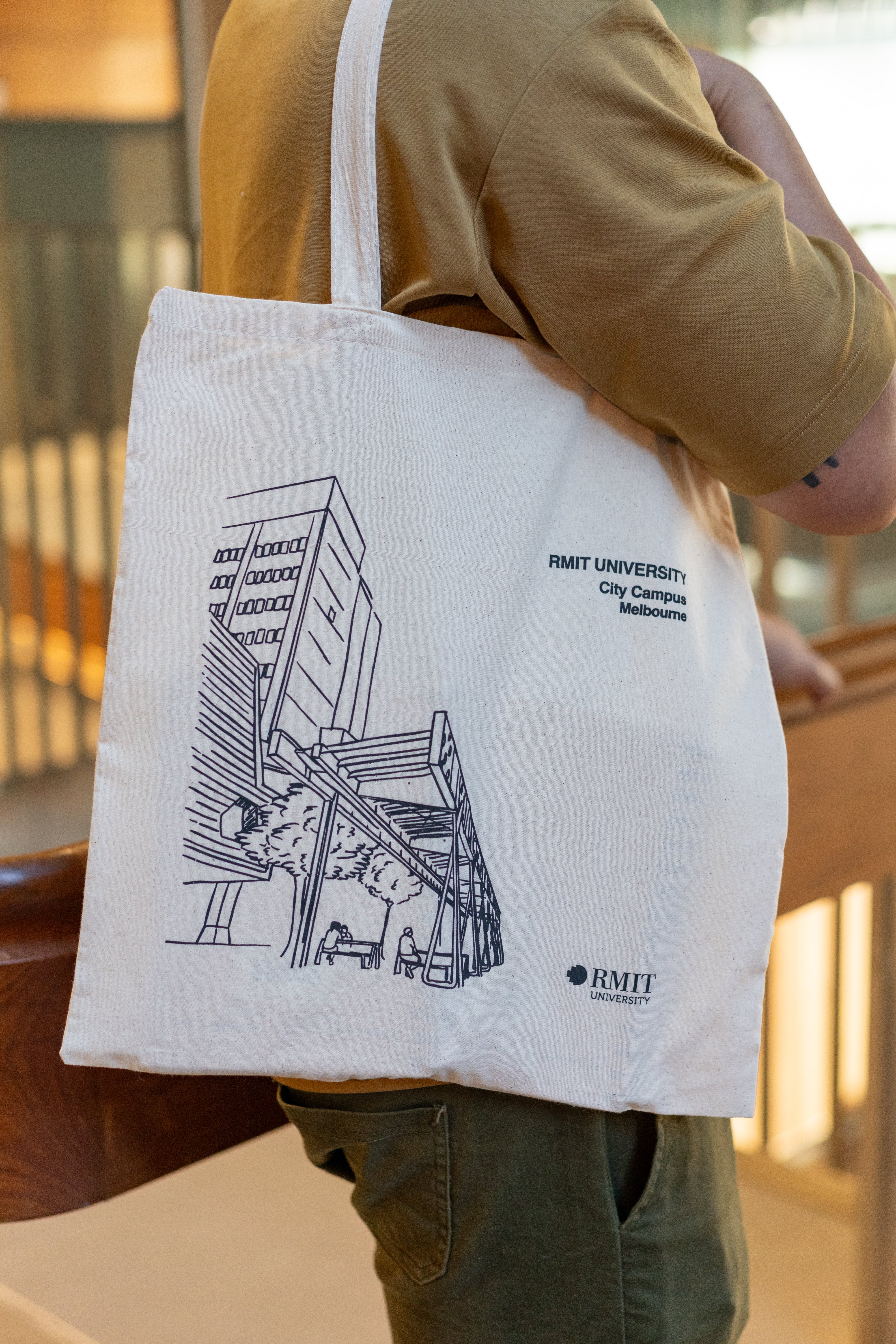 Side view of a model carrying the RMIT Store Everyday Campus Tote in natural cotton, featuring a detailed black line art illustration of the RMIT City Campus. The artwork includes the iconic Swanston Academic Building with its angular architecture and surrounding street-level details. The RMIT University logo and “City Campus” are printed above the illustration in black. The model wears a tan polo shirt and olive green cargo pants, photographed indoors with warm lighting.