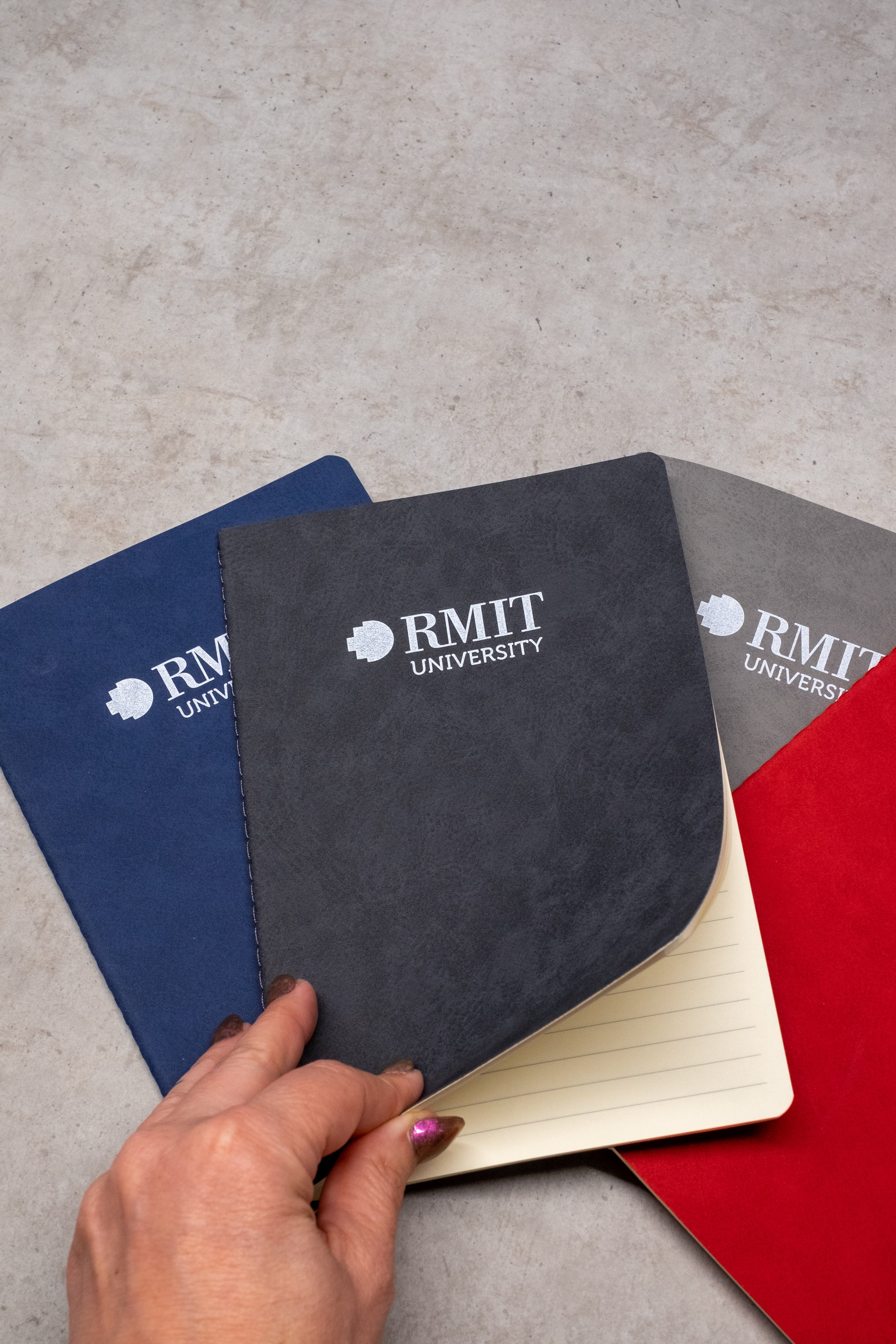 Hand opening a black Everyday Notebook from the RMIT Store, with blue and red notebooks in view, each featuring the RMIT University logo on the cover.