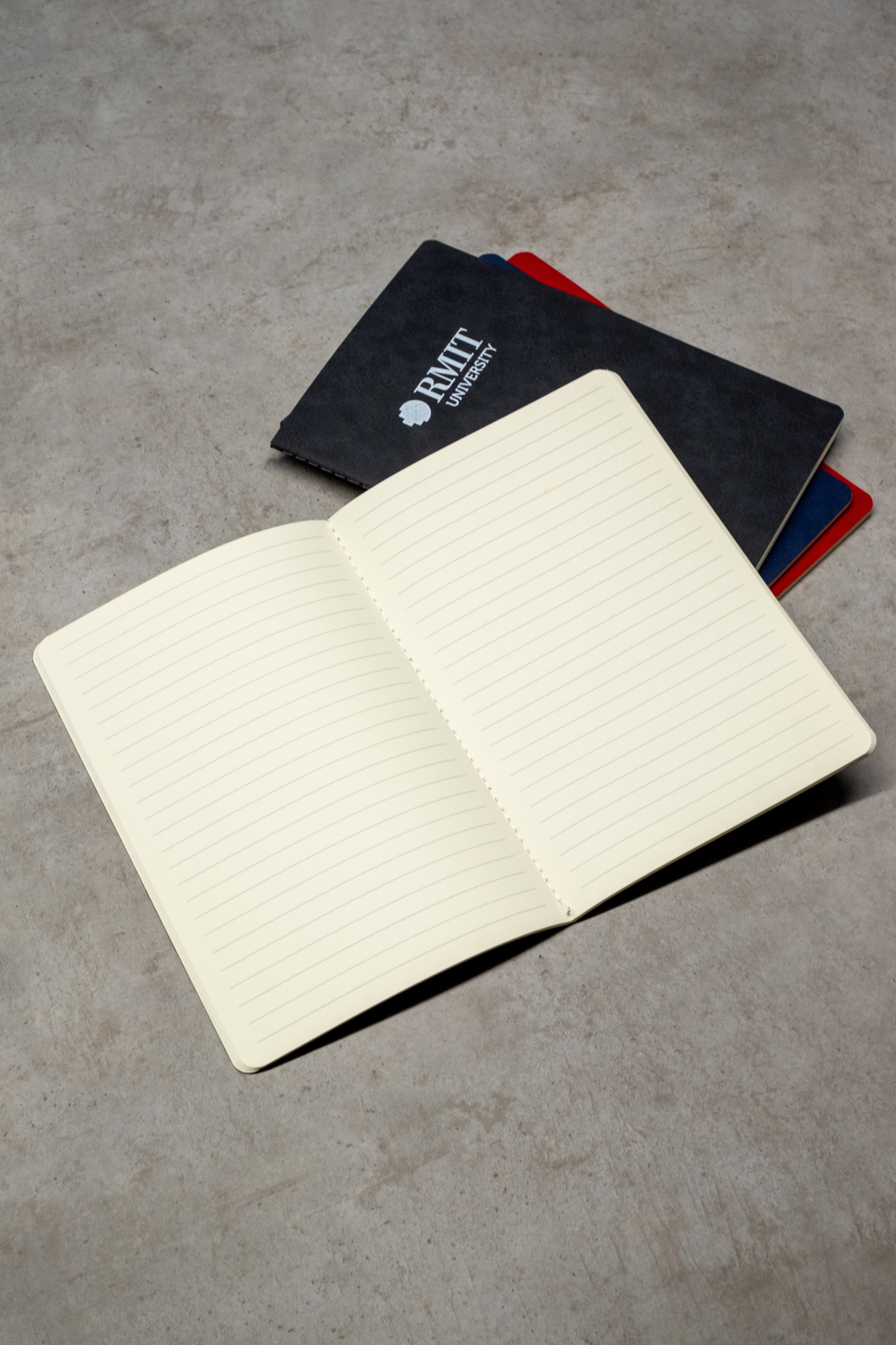 RMIT Store Everyday Notebook open to lined pages, with additional closed notebooks in black, blue, and red covers visible in the background.