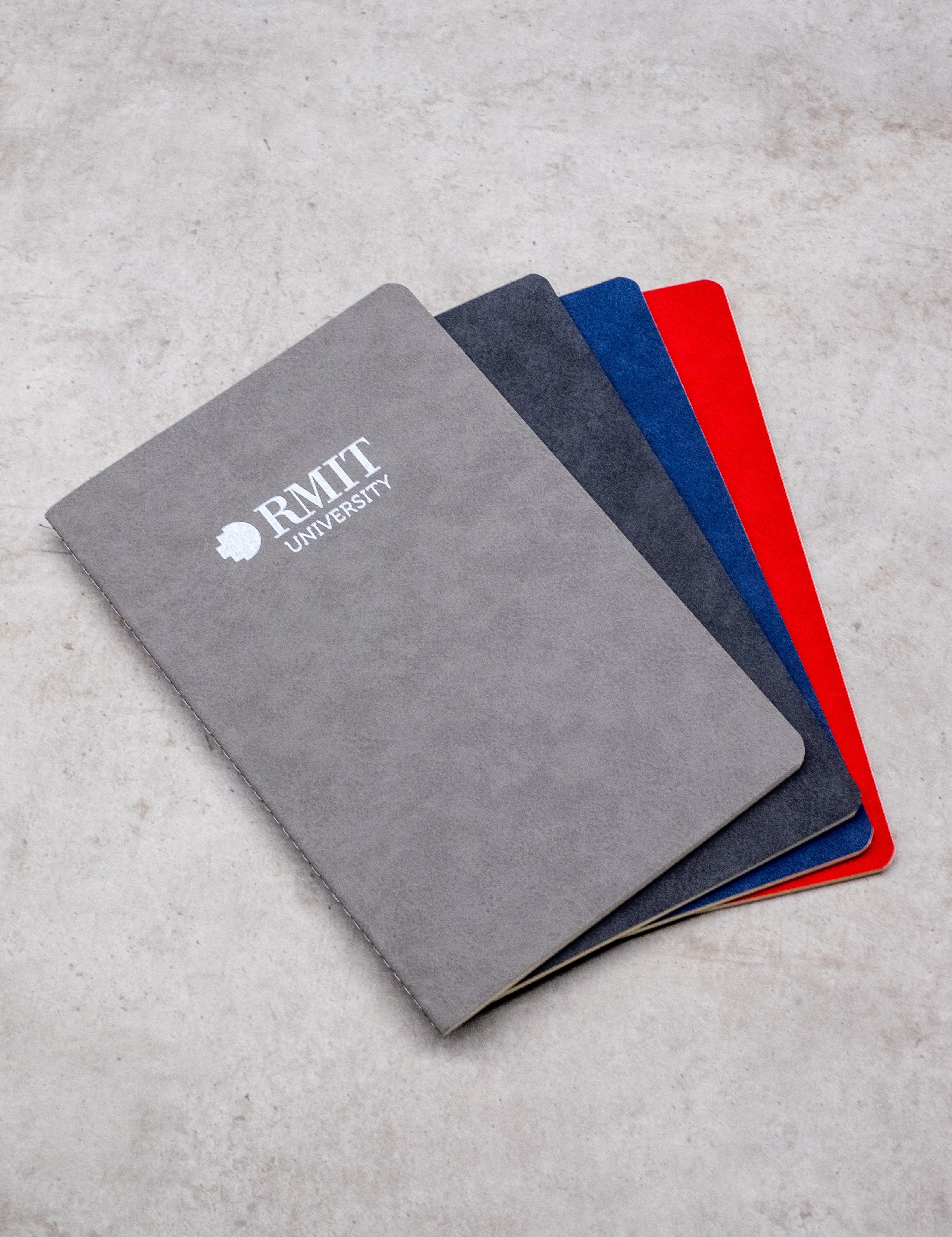 Set of Everyday Notebooks from the RMIT Store in grey, black, blue, and red covers, neatly stacked and showcasing the RMIT University logo.
