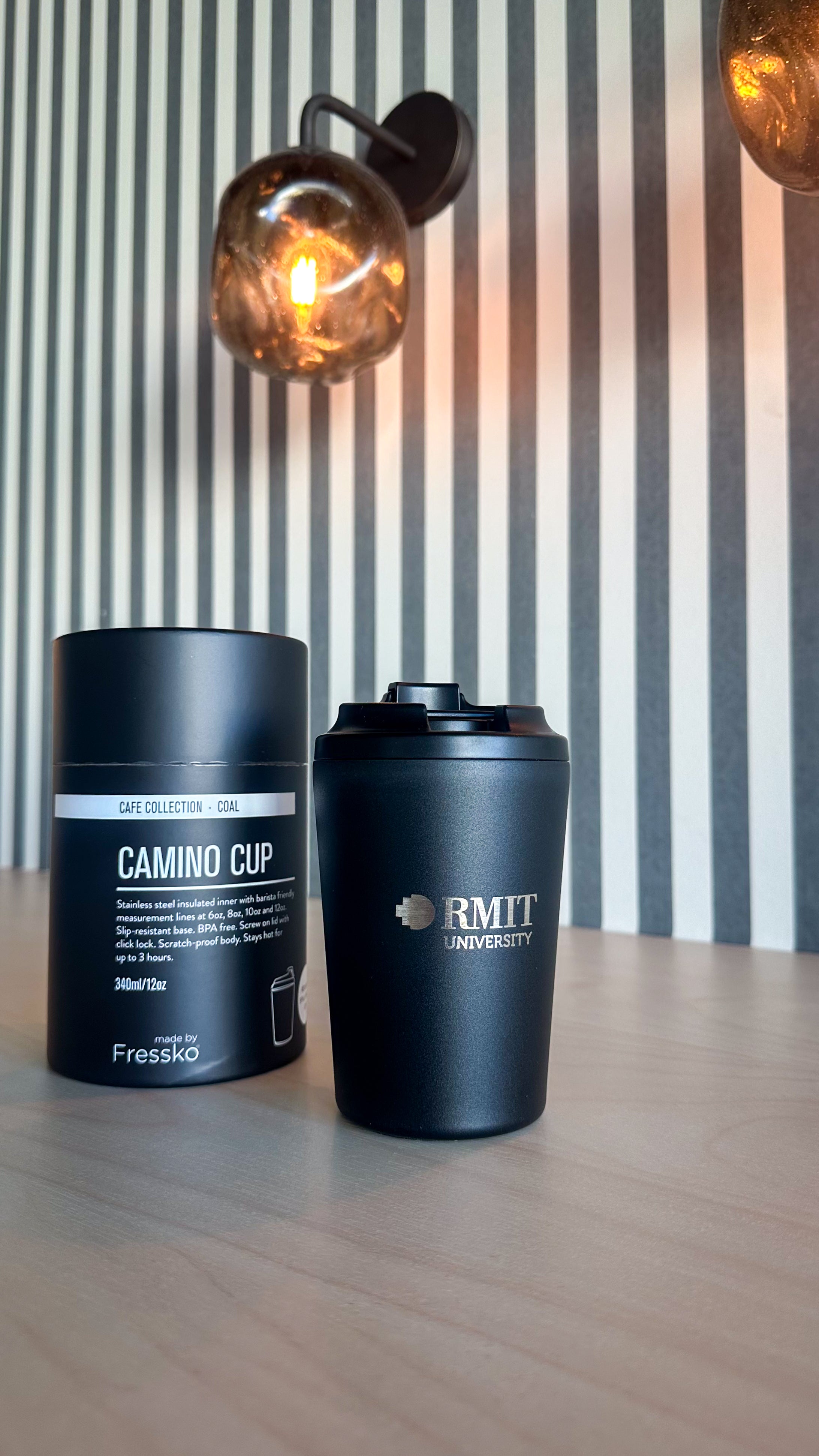 Coal Everyday Reusable Cup by madebyFressko in Coal with a laser-engraved silver RMIT University logo on the front, styled on a light timber table beside matching Camino Cup Coal packaging with warm pendant lighting and striped wall background, available at the RMIT Store.