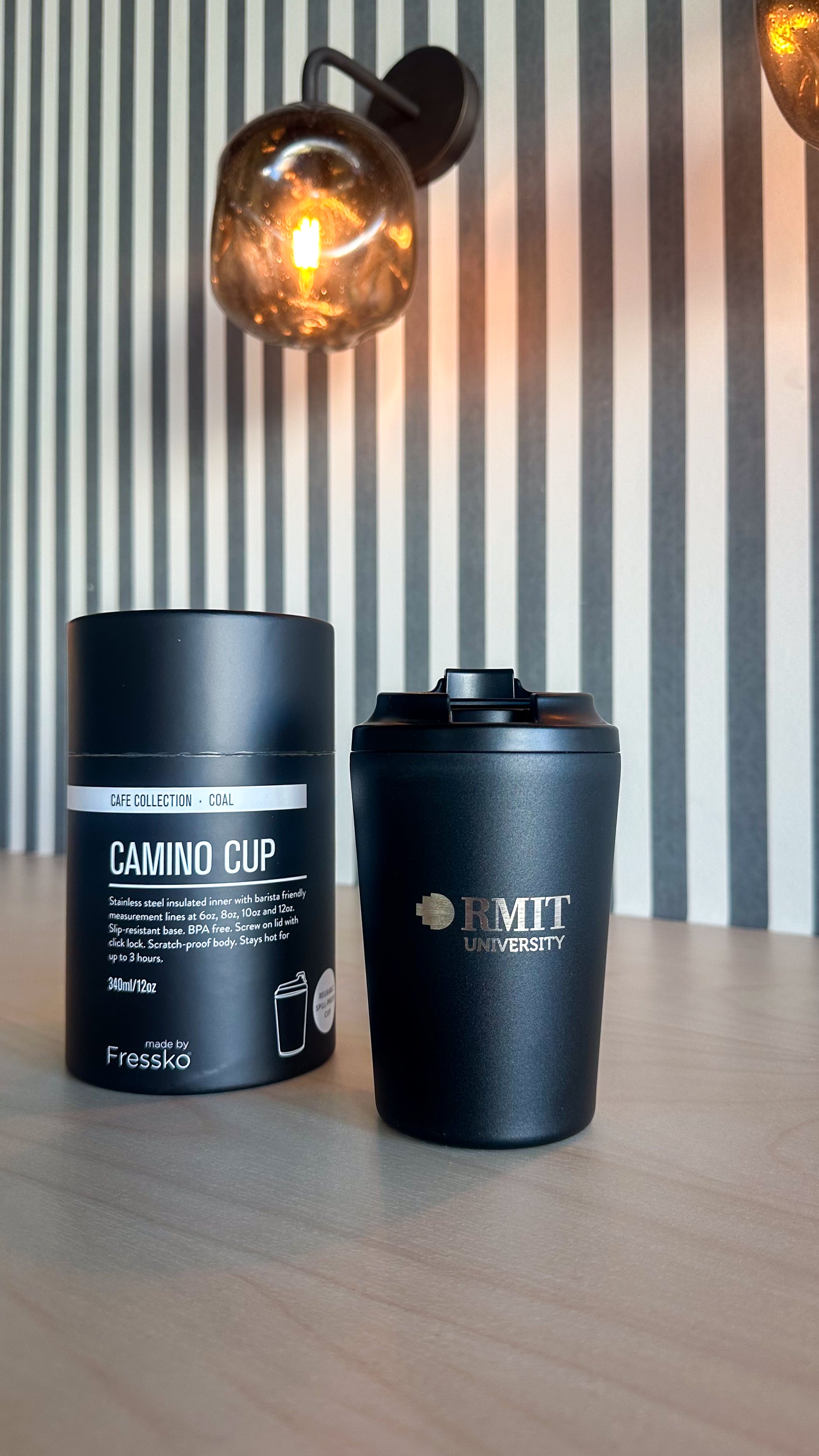 Coal Everyday Reusable Cup by madebyFressko in Coal with a laser-engraved silver RMIT University logo on the front, styled on a light timber table beside matching Camino Cup Coal packaging with warm pendant lighting and striped wall background, available at the RMIT Store.