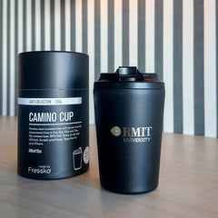 Coal Everyday Reusable Cup by madebyFressko in Coal with a laser-engraved silver RMIT University logo on the front, styled on a light timber table beside matching Camino Cup Coal packaging with warm pendant lighting and striped wall background, available at the RMIT Store.