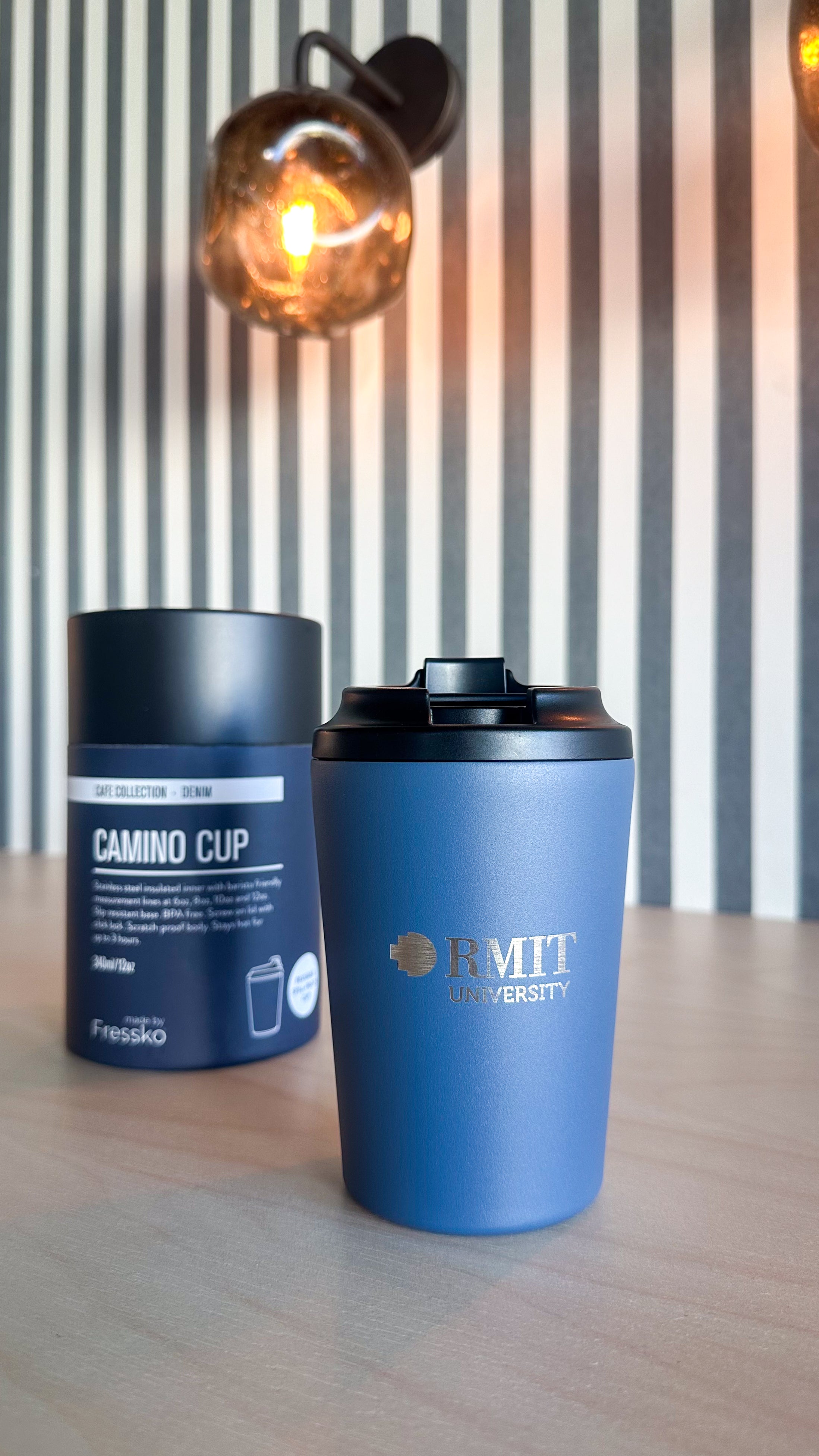 Denim Everyday Reusable Cup by madebyFressko in muted denim blue with an engraved silver RMIT University logo on the front, displayed on a light timber tabletop with matching Camino Cup Denim packaging softly blurred in the background against a striped wall and warm ambient lighting, available at the RMIT Store.