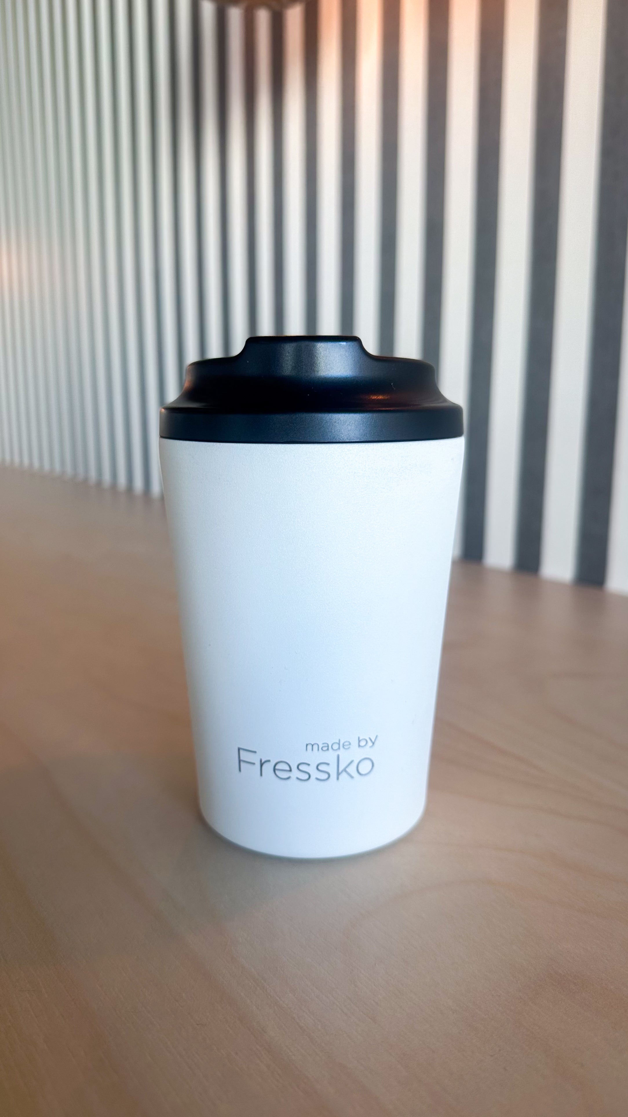 Frost Everyday Reusable Cup by madebyFressko in matte frost white with subtle grey made by Fressko branding near the base and a black screw-top lid, photographed on a light timber table against a striped wall background with soft warm lighting, available at the RMIT Store.