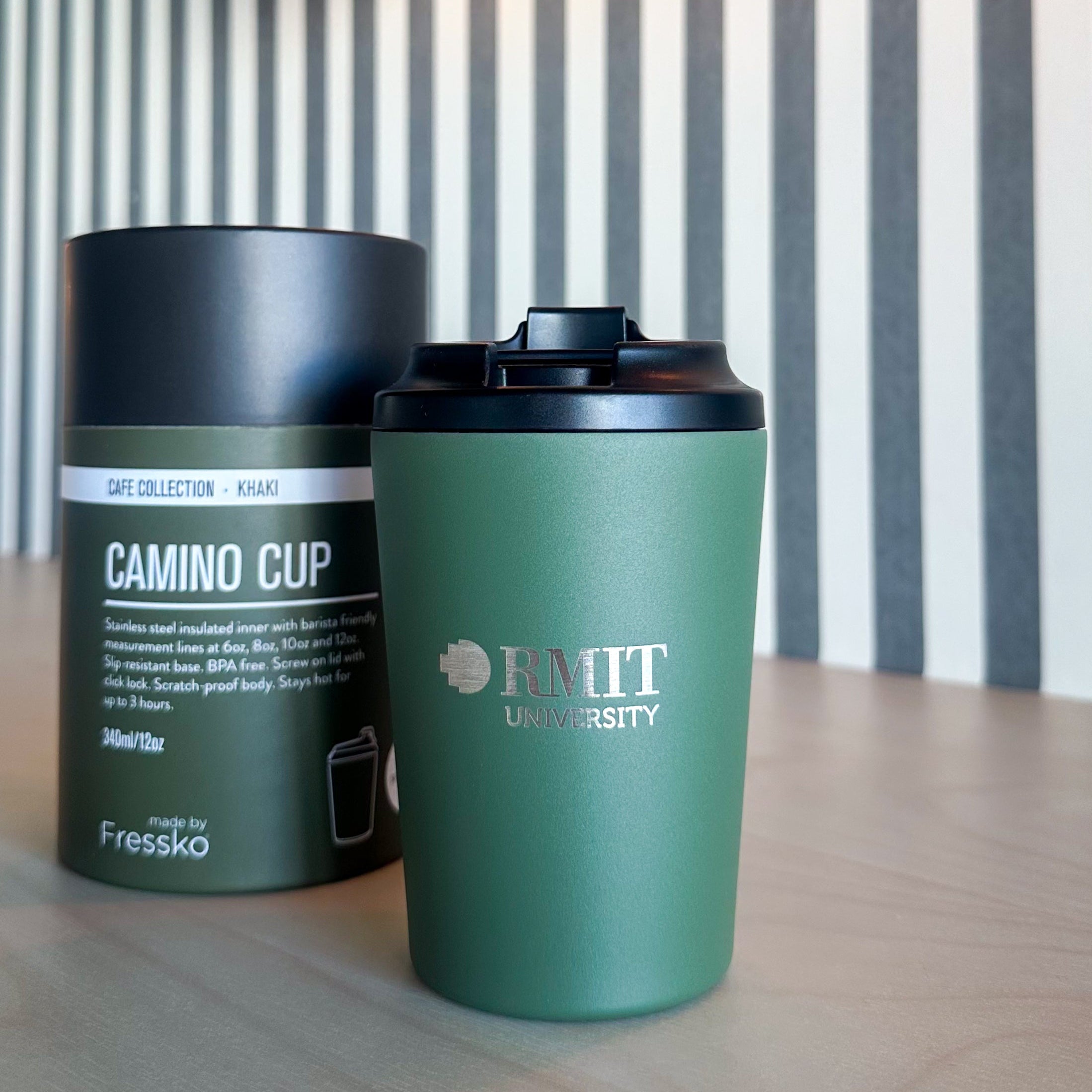 Everyday Reusable Cup in Khaki with a matte green finish, black screw-on sip lid and silver RMIT University logo on the front, displayed beside its cylindrical Fressko Café Collection Khaki packaging on a timber table, with Denim, Frost and Black RMIT University reusable cups and packaging softly out of focus in the background at RMIT Store.