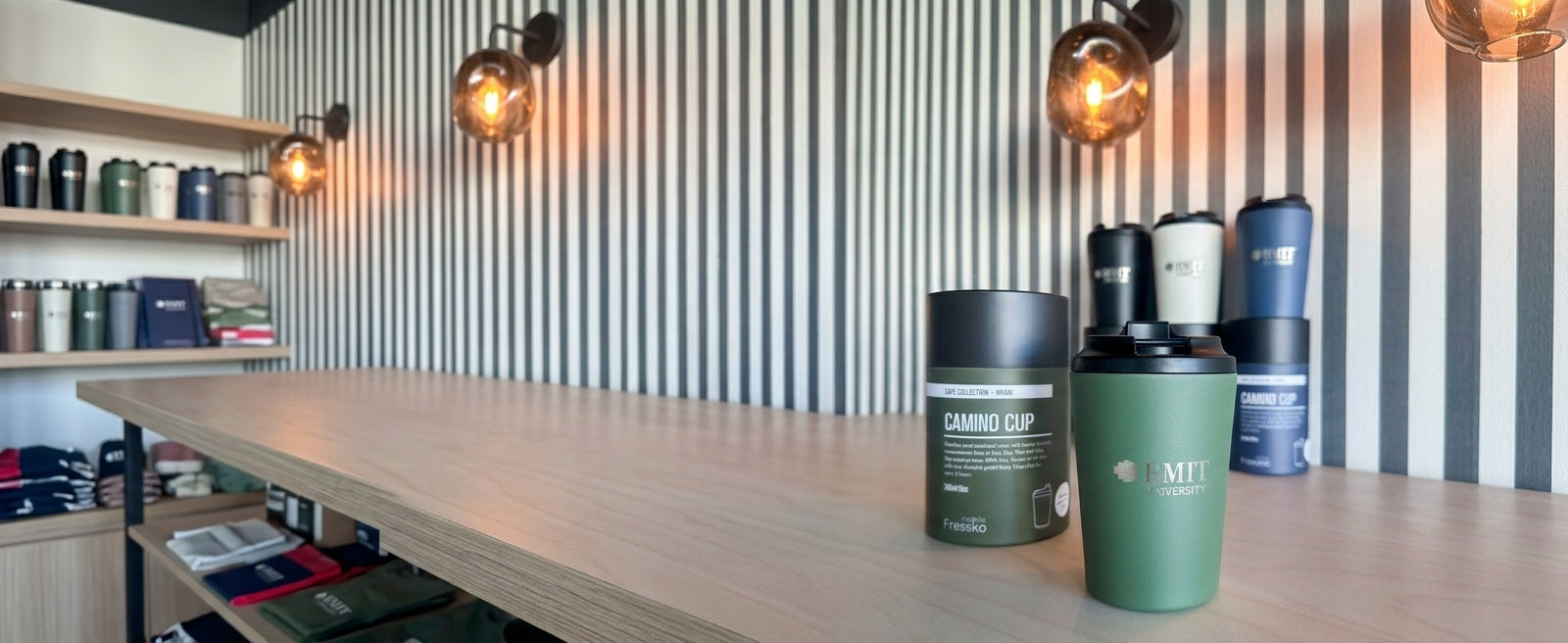 Everyday Reusable Cup in Khaki with a matte green finish, black screw-on sip lid and silver RMIT University logo on the front, displayed beside its cylindrical Fressko Café Collection Khaki packaging on a timber table, with Denim, Frost and Black RMIT University reusable cups and packaging softly out of focus in the background at RMIT Store.