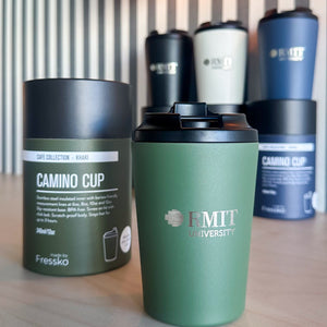 Everyday Reusable Cup in Khaki with a matte green finish, black screw-on sip lid and silver RMIT University logo on the front, displayed beside its cylindrical Fressko Café Collection Khaki packaging on a timber table, with striped wall panelling and a warm glass wall light in the background at RMIT Store.