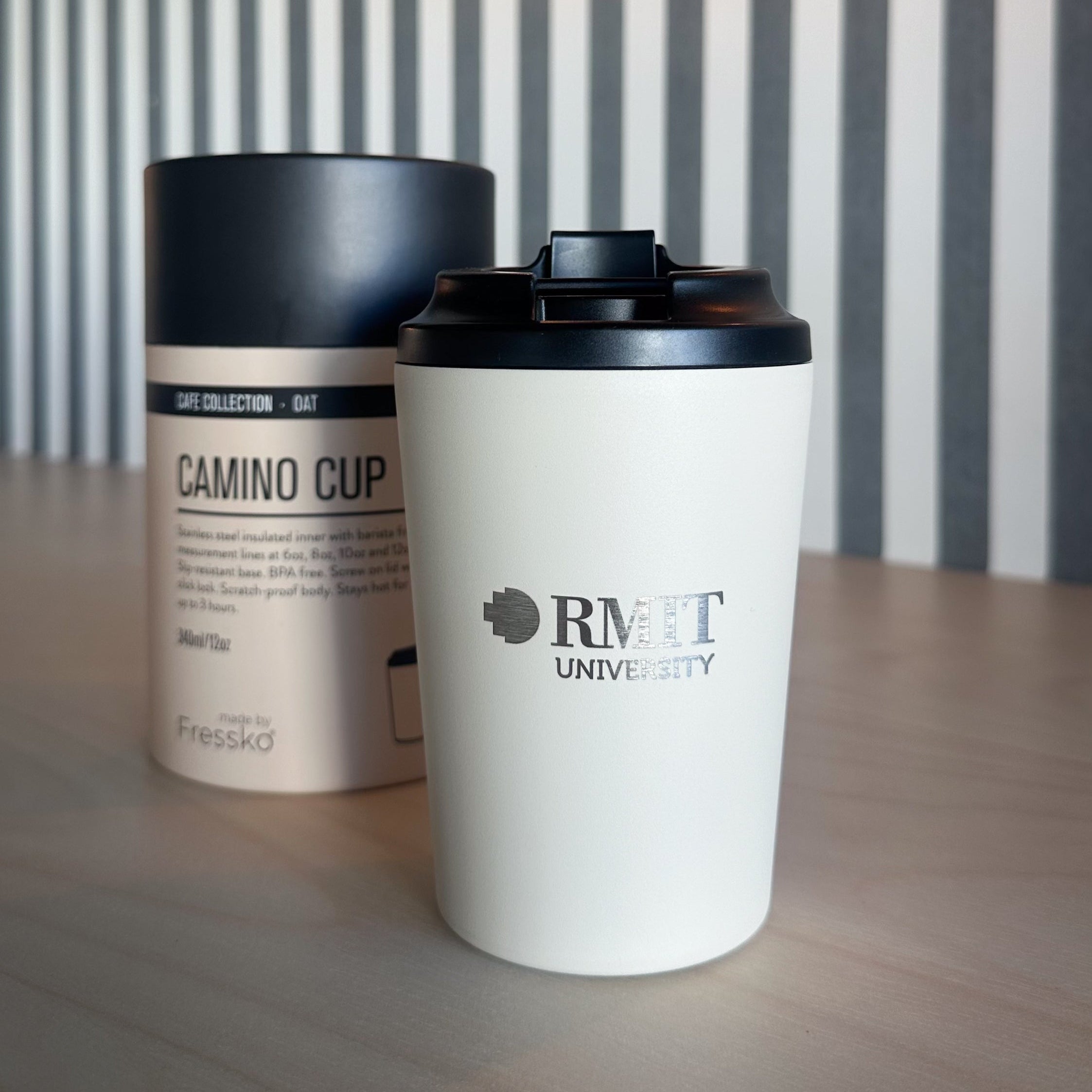 Everyday Reusable Cup in Oat, standing upright on a light timber table with a matte off-white body, black flip-top lid and silver RMIT University logo printed on the front, positioned beside its matching Oat Fressko Camino Cup packaging against a grey-and-white striped wall with a warm globe light for the RMIT Store.
