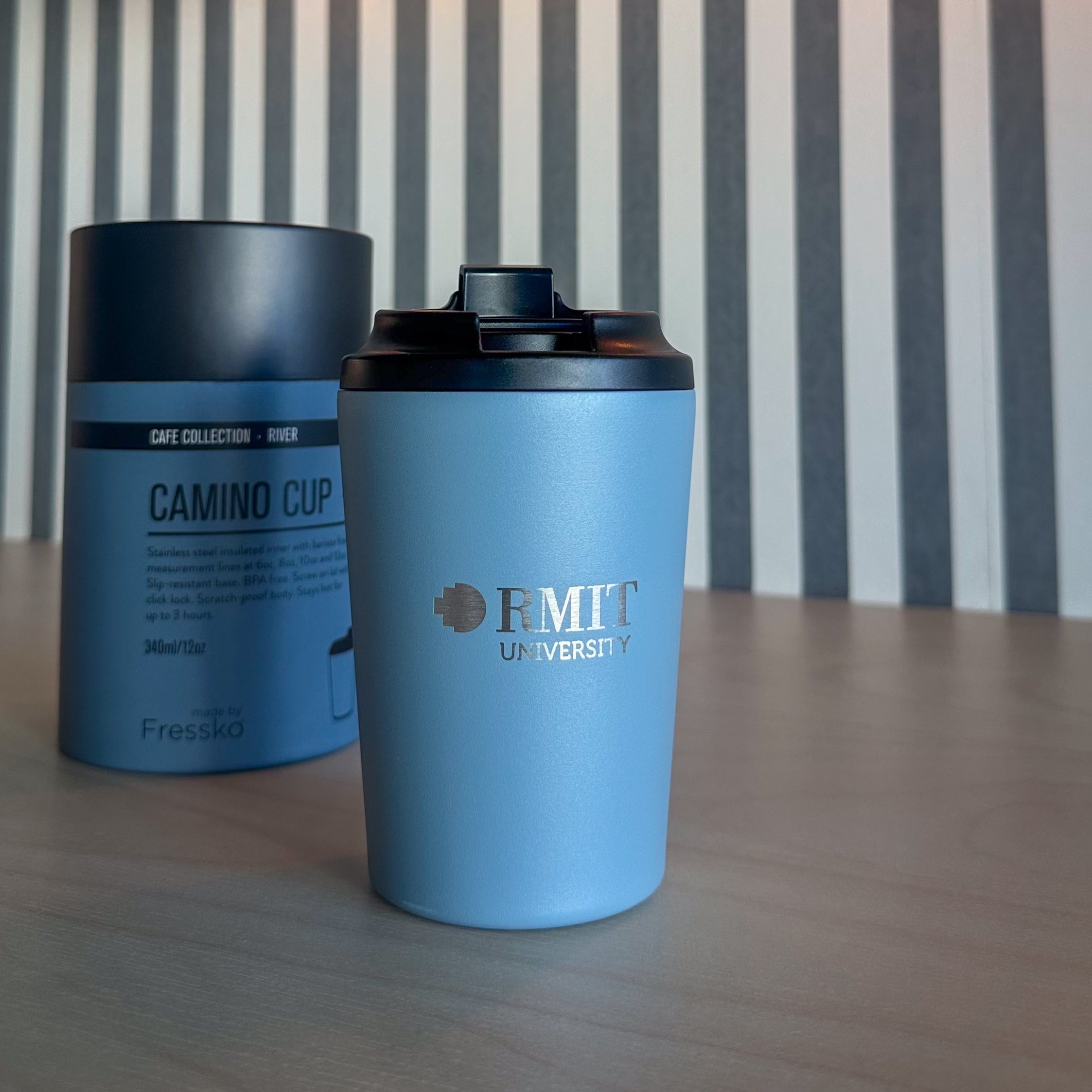 Everyday Reusable Cup in River blue, standing upright on a light timber table with a matte blue body, black flip-top lid and silver RMIT University logo printed on the front, positioned beside its matching blue Fressko Camino Cup packaging against a grey-and-white striped wall with warm globe lights for the RMIT Store.