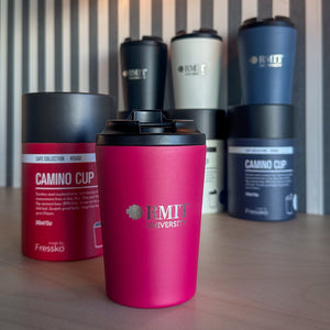 RMIT Store Everyday Reusable Cup in Rouge, shown from the front with a matte finish, black click-lock lid and silver RMIT University logo, displayed on a timber table with Coal, Frost and Denim reusable cups and matching Fressko packaging softly blurred in the background.