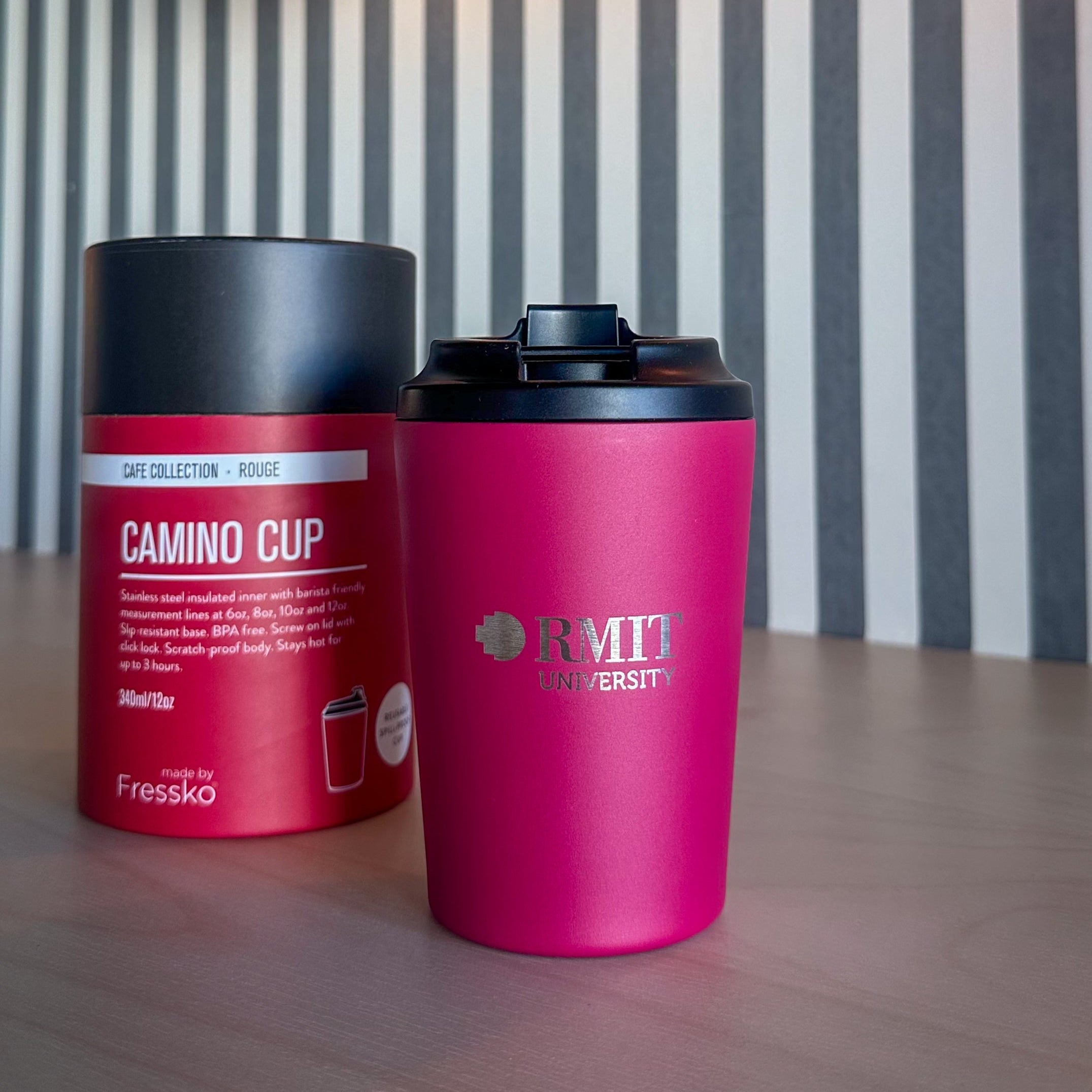 RMIT Store Everyday Reusable Cup in Rouge, shown from the front with a matte finish, black click-lock lid and silver RMIT University logo, displayed beside its cylindrical Fressko Camino Cup packaging labelled Café Collection Rouge and 340 ml on a timber table beneath an amber glass light.