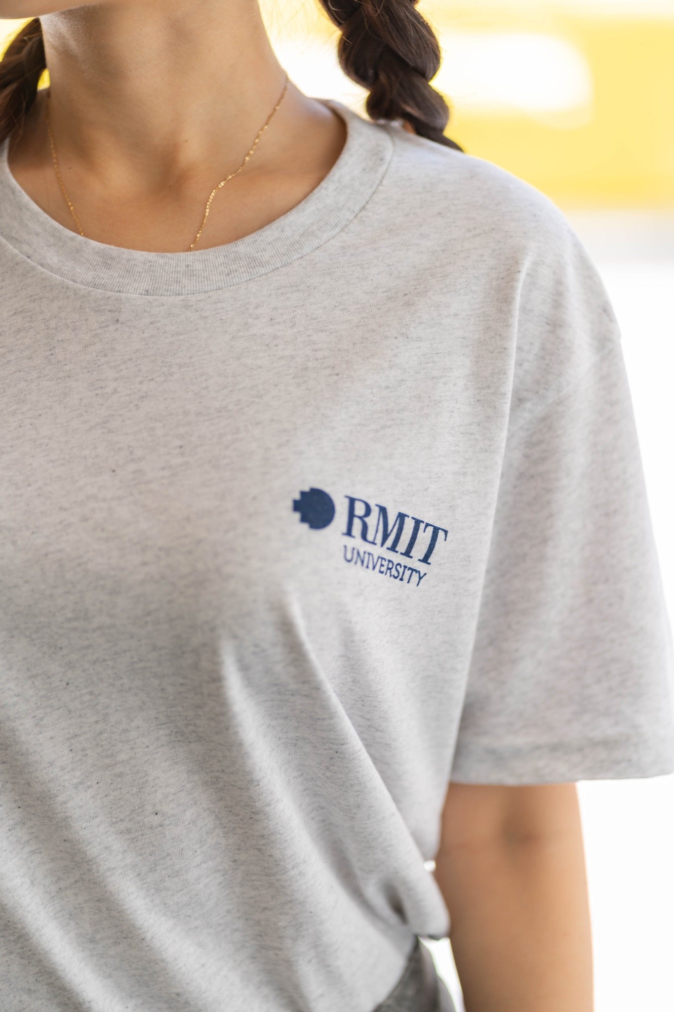 Everyday Tee in white marle, close-up front view worn by a model showing the upper torso and neckline, featuring a soft heathered cotton fabric with subtle grey flecks, a small navy RMIT University logo printed on the left chest, crew neckline and smooth stitching, styled outdoors with warm natural lighting and blurred background, available at the RMIT Store.