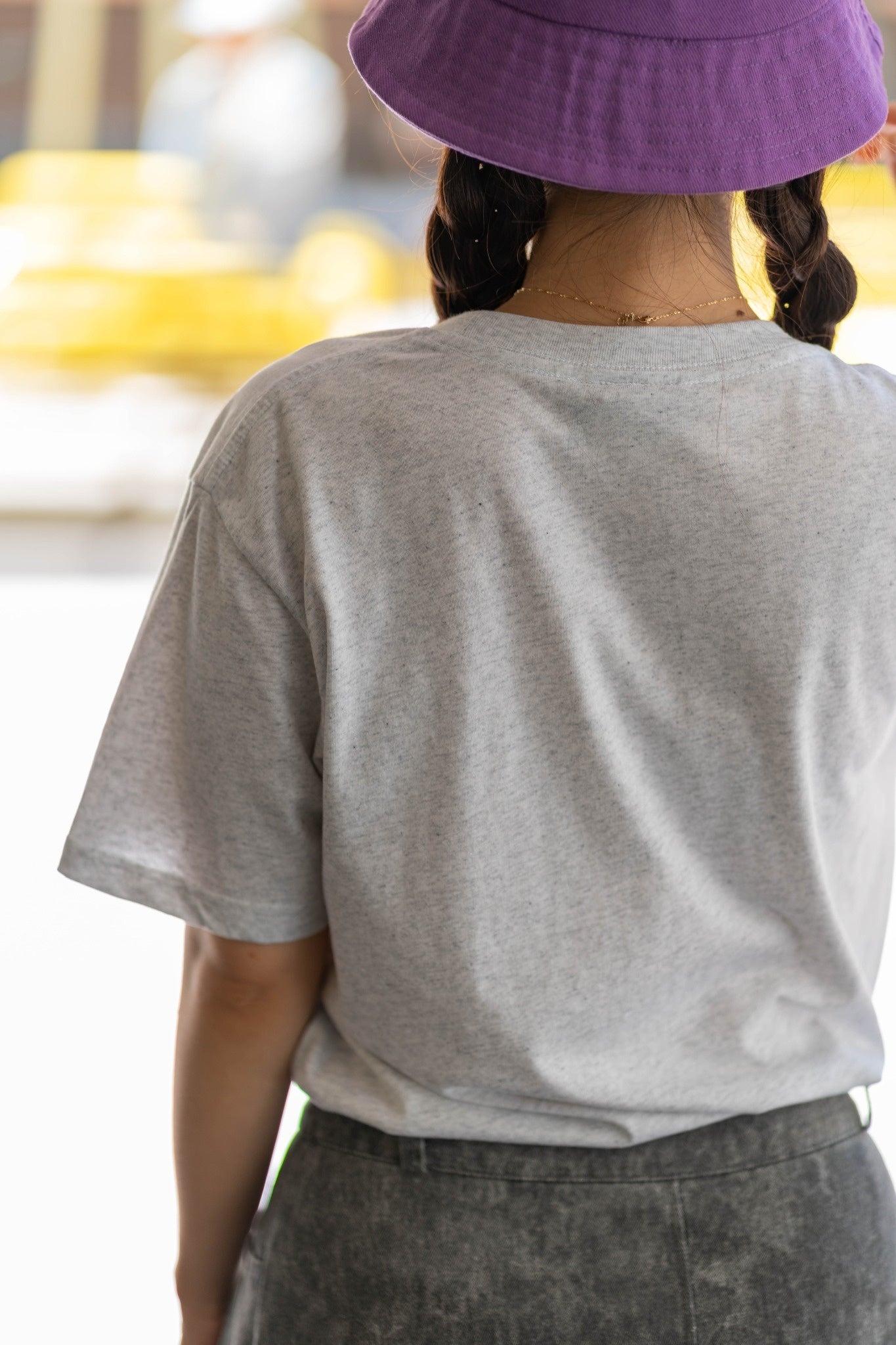 Everyday Tee in white marle, rear view worn by a model standing outdoors, featuring a plain back with no visible branding, soft heathered cotton fabric with fine grey flecks, relaxed fit with short sleeves and crew neckline, styled with a purple bucket hat and braided hair against a softly blurred outdoor background, available at the RMIT Store.
