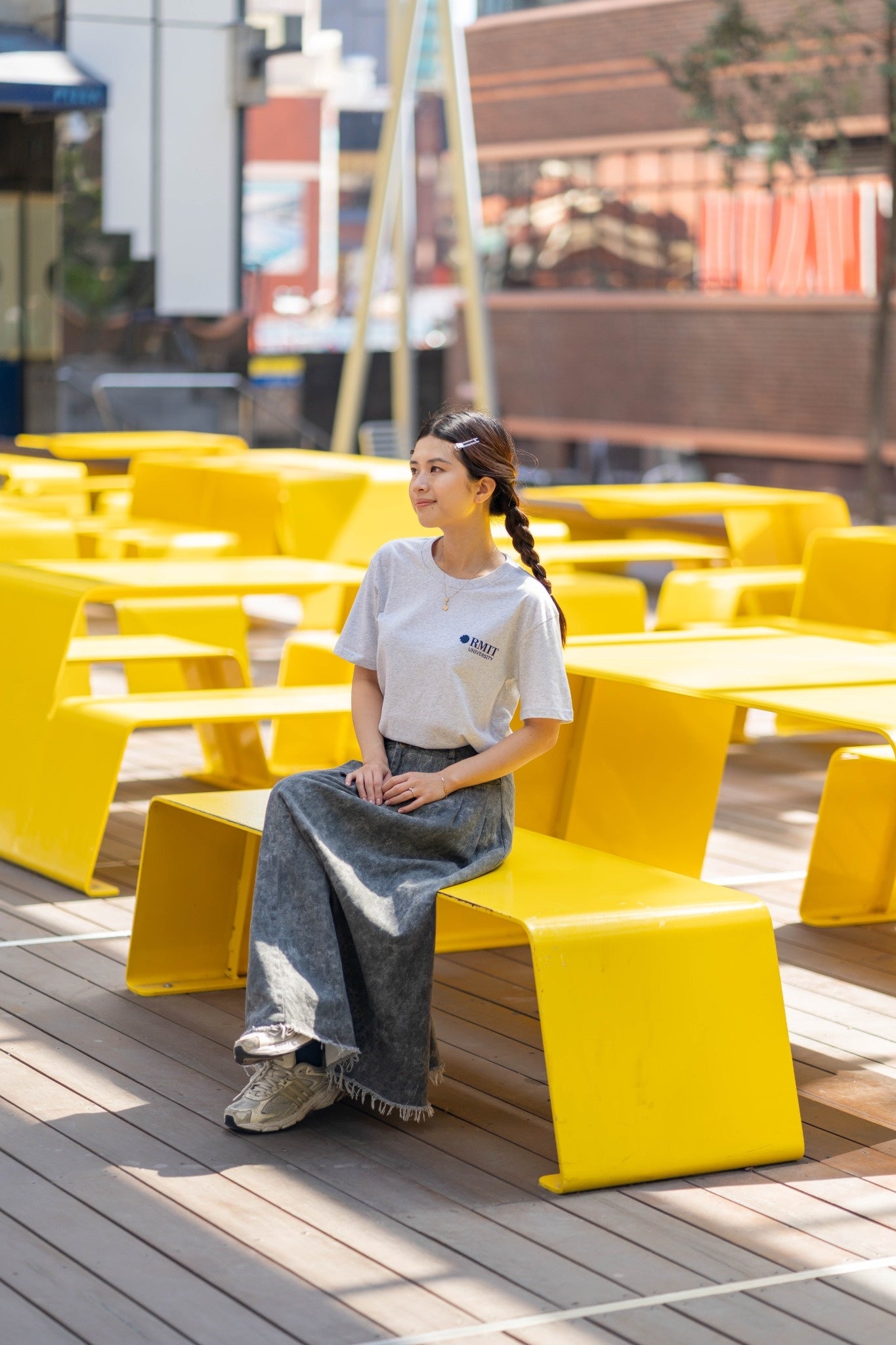 Everyday Tee in white marle, full body lifestyle view worn by a seated model on bright yellow outdoor benches, featuring a small navy RMIT University logo on the left chest, soft heathered cotton fabric with grey flecks, relaxed fit paired with a long grey skirt and sneakers, set in a sunlit urban courtyard with multiple yellow benches, available at the RMIT Store.