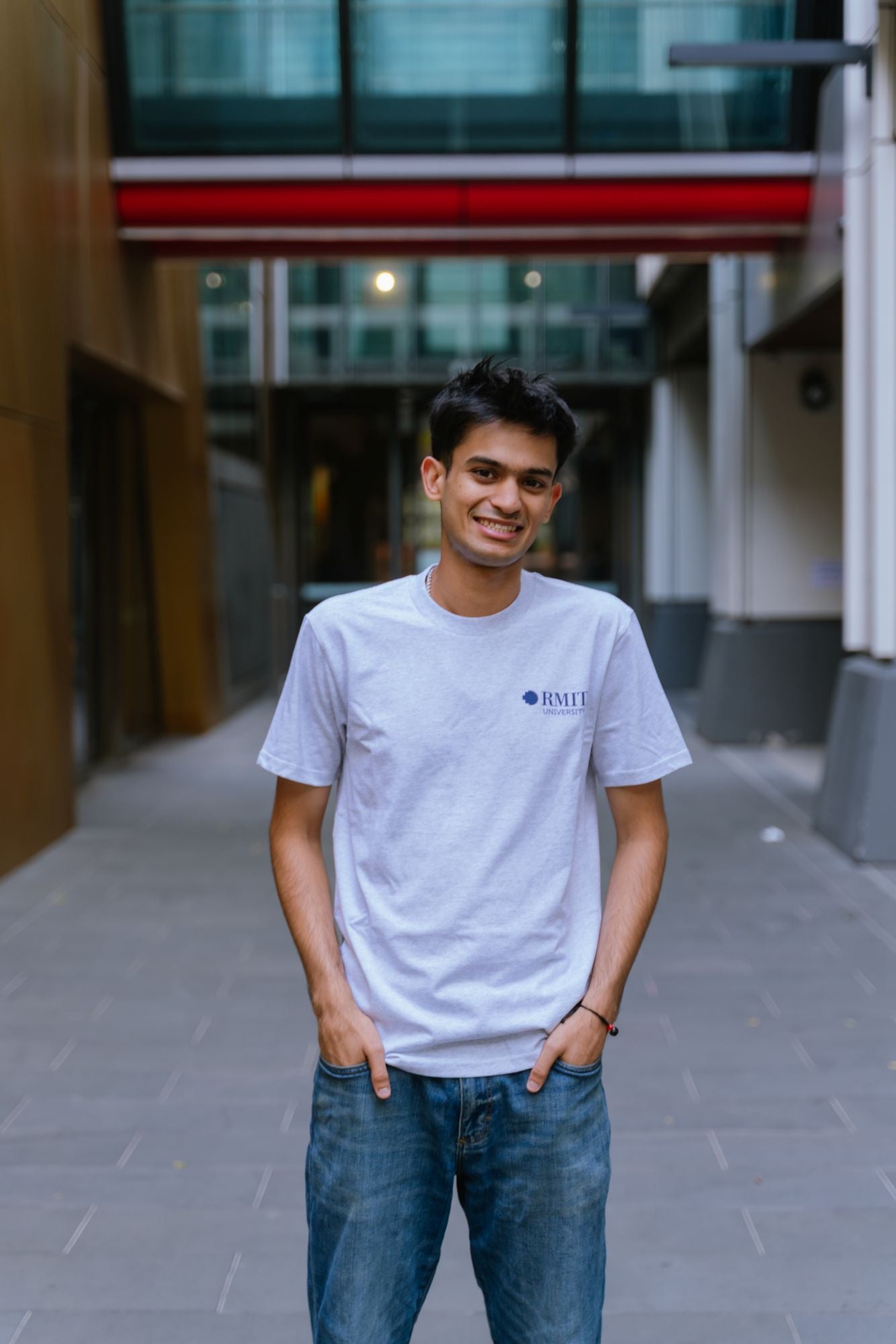 Model standing in an outdoor campus walkway wearing Everyday Tee in White Marle with a small RMIT University logo printed on the left chest, paired with blue denim jeans, hands in pockets and smiling, with blurred modern building background, available at the RMIT Store.
