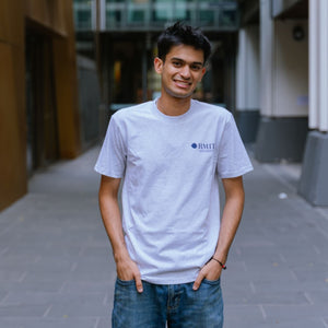 Model standing in an outdoor campus walkway wearing Everyday Tee in White Marle with a small RMIT University logo printed on the left chest, paired with blue denim jeans, hands in pockets and smiling, with blurred modern building background, available at the RMIT Store.