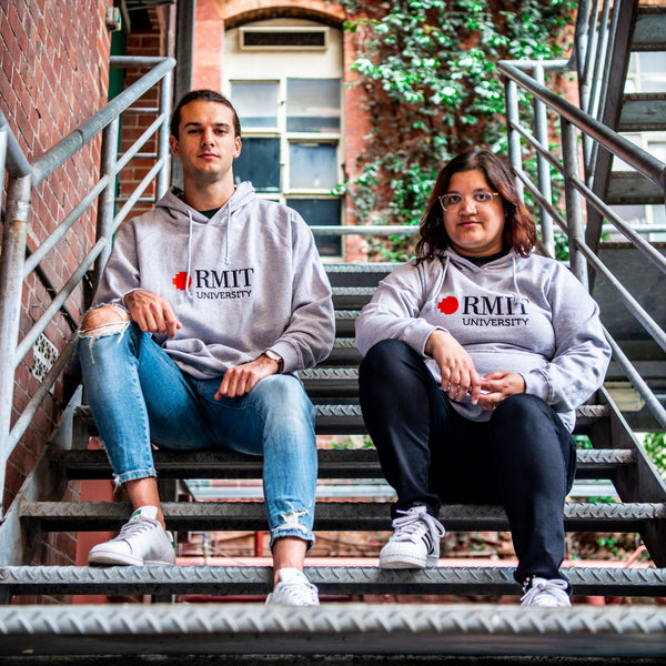 Two models seated on an outdoor metal staircase wearing Fairtrade Hoodie in grey with a front-printed RMIT University logo featuring a red pixel square and black text, showcasing a relaxed fit cotton fleece design with ribbed cuffs and drawstring hood, styled with casual denim and black trousers against a red brick building with green ivy background, available at the RMIT Store.