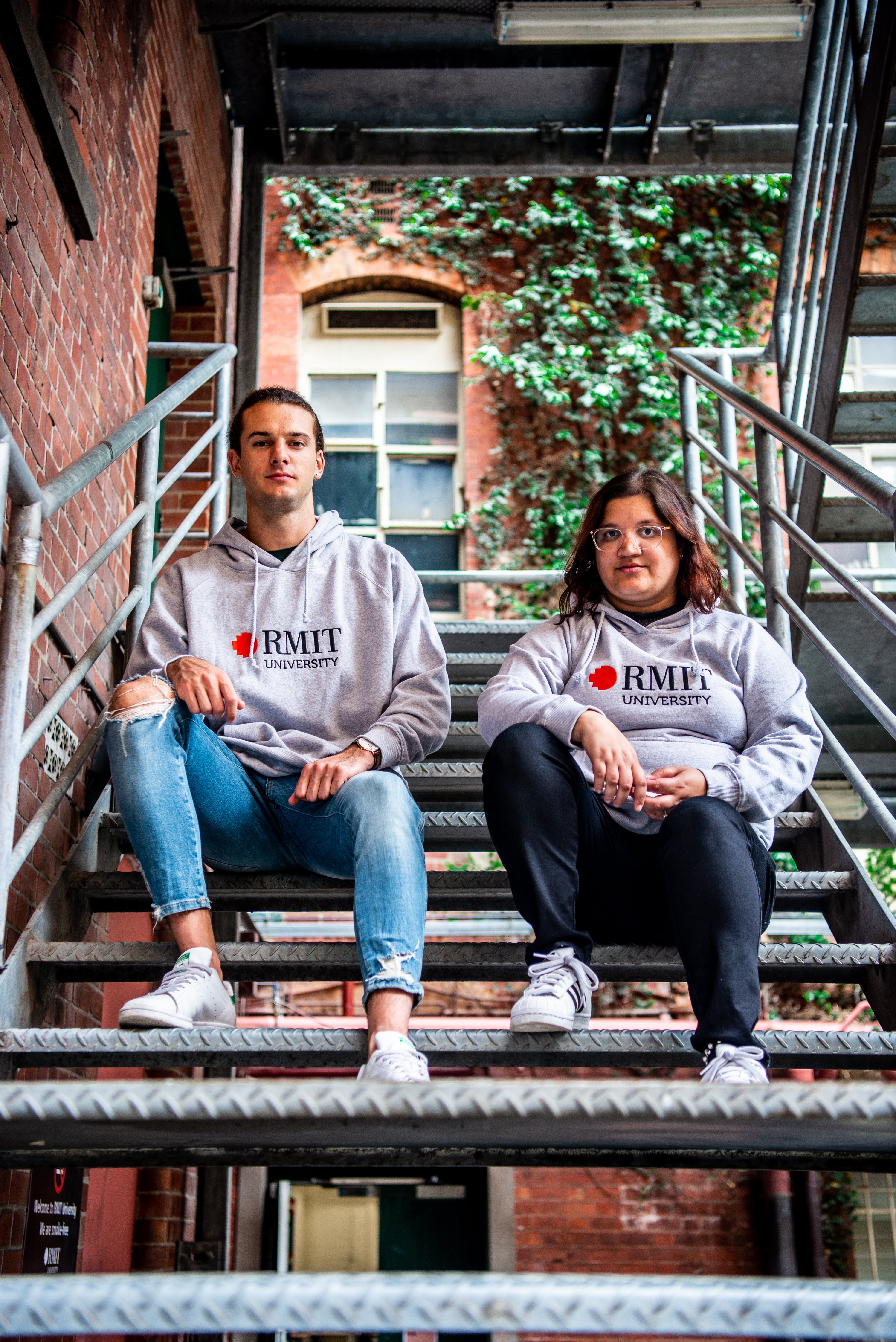 Two models seated on an outdoor metal staircase wearing Fairtrade Hoodie in grey with a front-printed RMIT University logo featuring a red pixel square and black text, showcasing a relaxed fit cotton fleece design with ribbed cuffs and drawstring hood, styled with casual denim and black trousers against a red brick building with green ivy background, available at the RMIT Store.