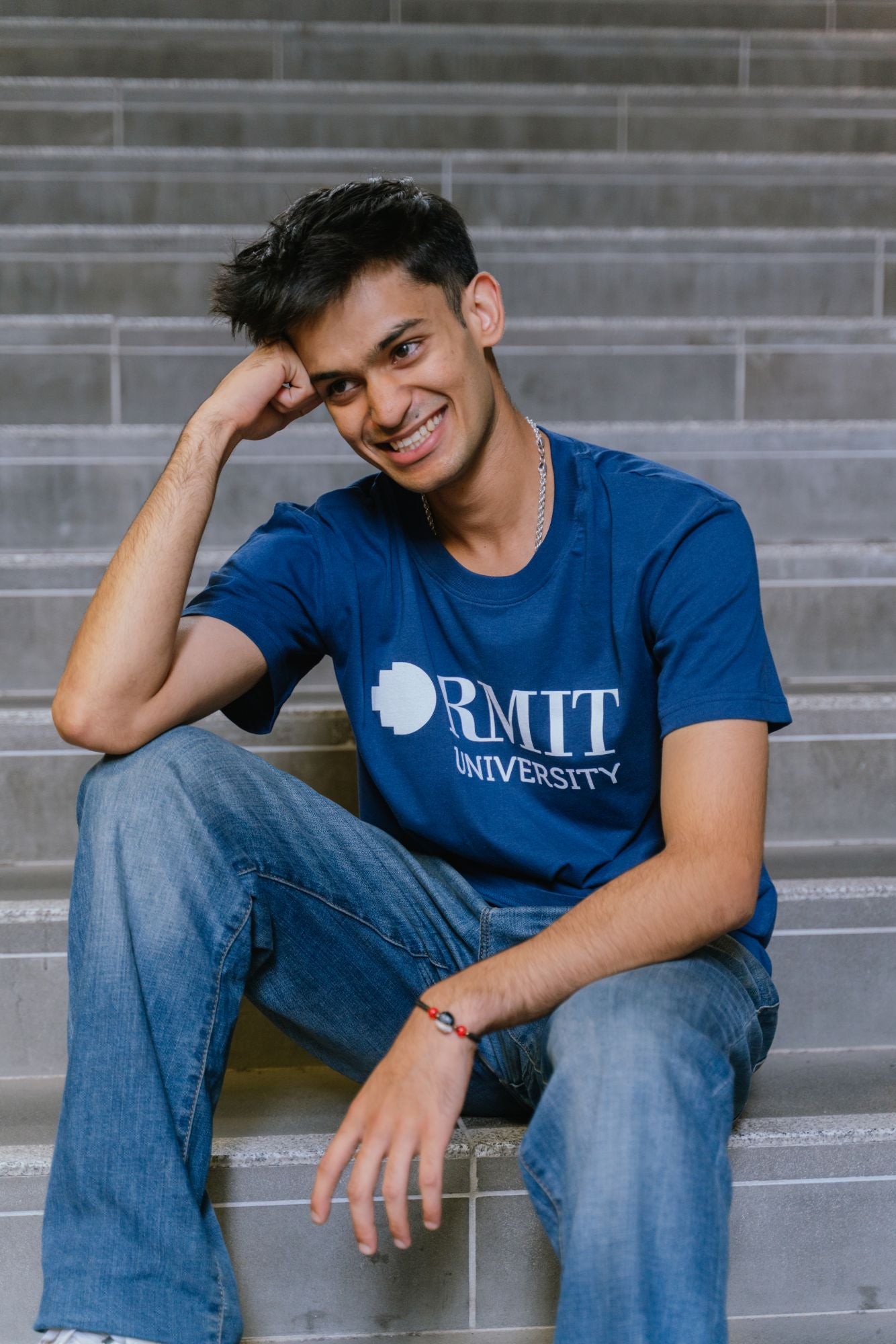 Model sitting on indoor steps wearing Fairtrade Loud Logo Tee in navy featuring a large white RMIT University logo across the chest, paired with blue denim jeans, leaning slightly forward with hands clasped, with soft lighting highlighting the printed logo detail, available at the RMIT Store.