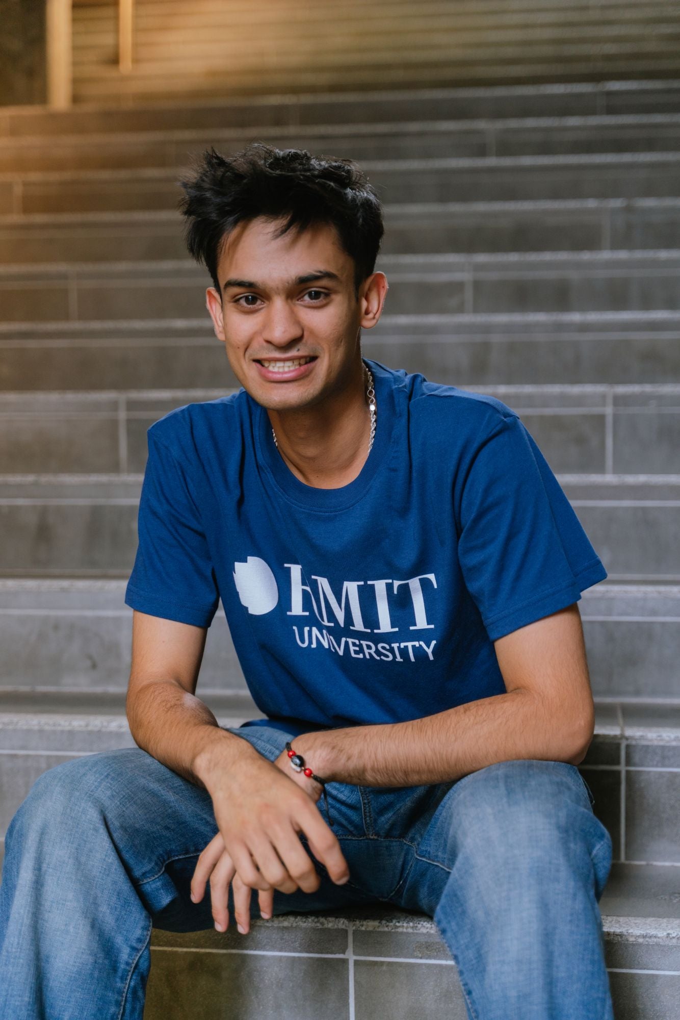 Model sitting on indoor concrete stairs wearing Fairtrade Loud Logo Tee in navy featuring a large white RMIT University logo printed prominently across the chest, paired with blue denim jeans, smiling with one arm resting on knee, in a softly lit architectural stairwell, available at the RMIT Store.