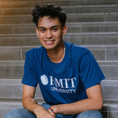 Model sitting on indoor concrete stairs wearing Fairtrade Loud Logo Tee in navy featuring a large white RMIT University logo printed prominently across the chest, paired with blue denim jeans, smiling with one arm resting on knee, in a softly lit architectural stairwell, available at the RMIT Store.