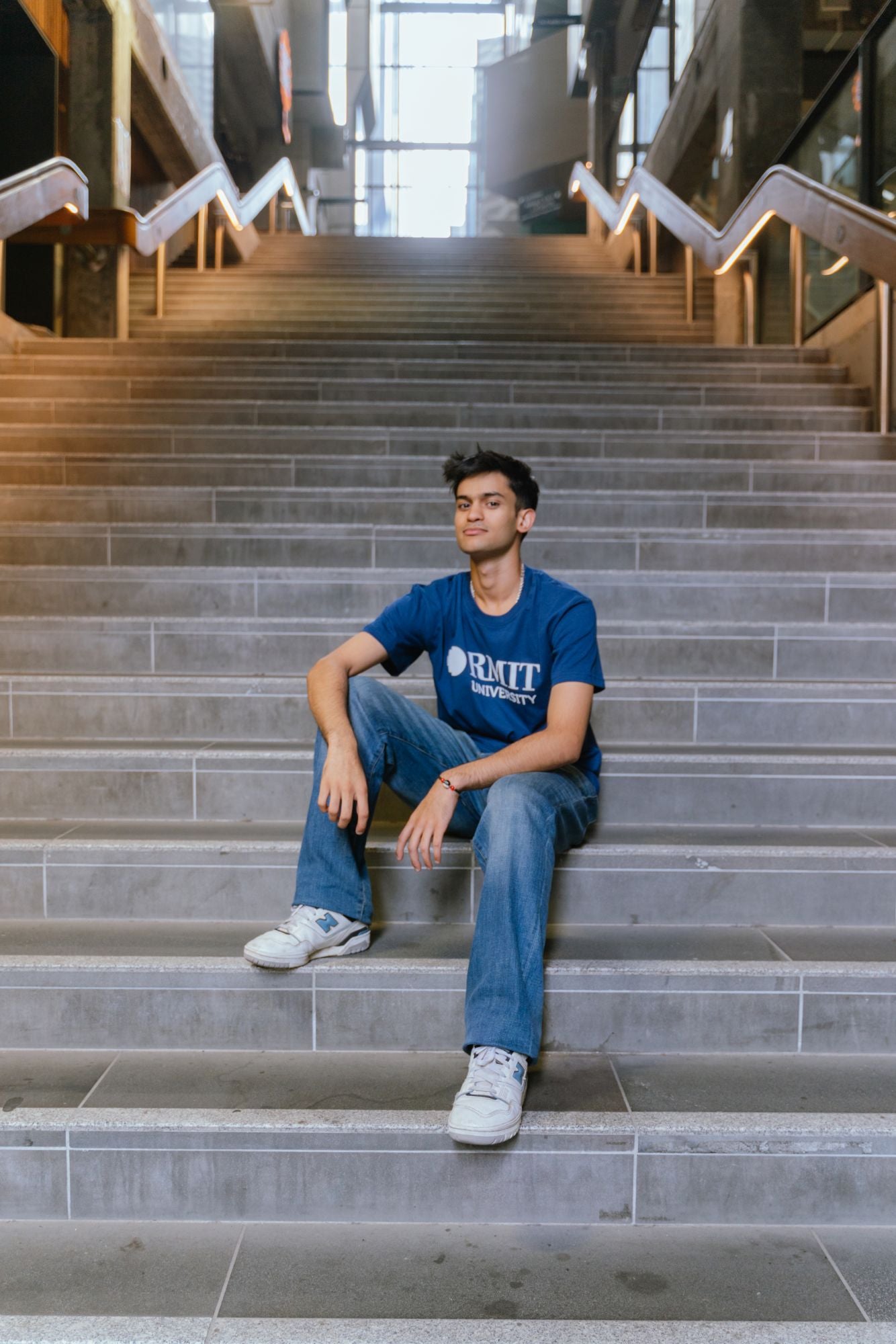 Model seated centrally on wide indoor concrete stairs wearing Fairtrade Loud Logo Tee in navy with bold white RMIT University logo across the chest, styled with blue denim jeans and white sneakers, relaxed pose with hands resting on legs in a modern stairwell setting, available at the RMIT Store.