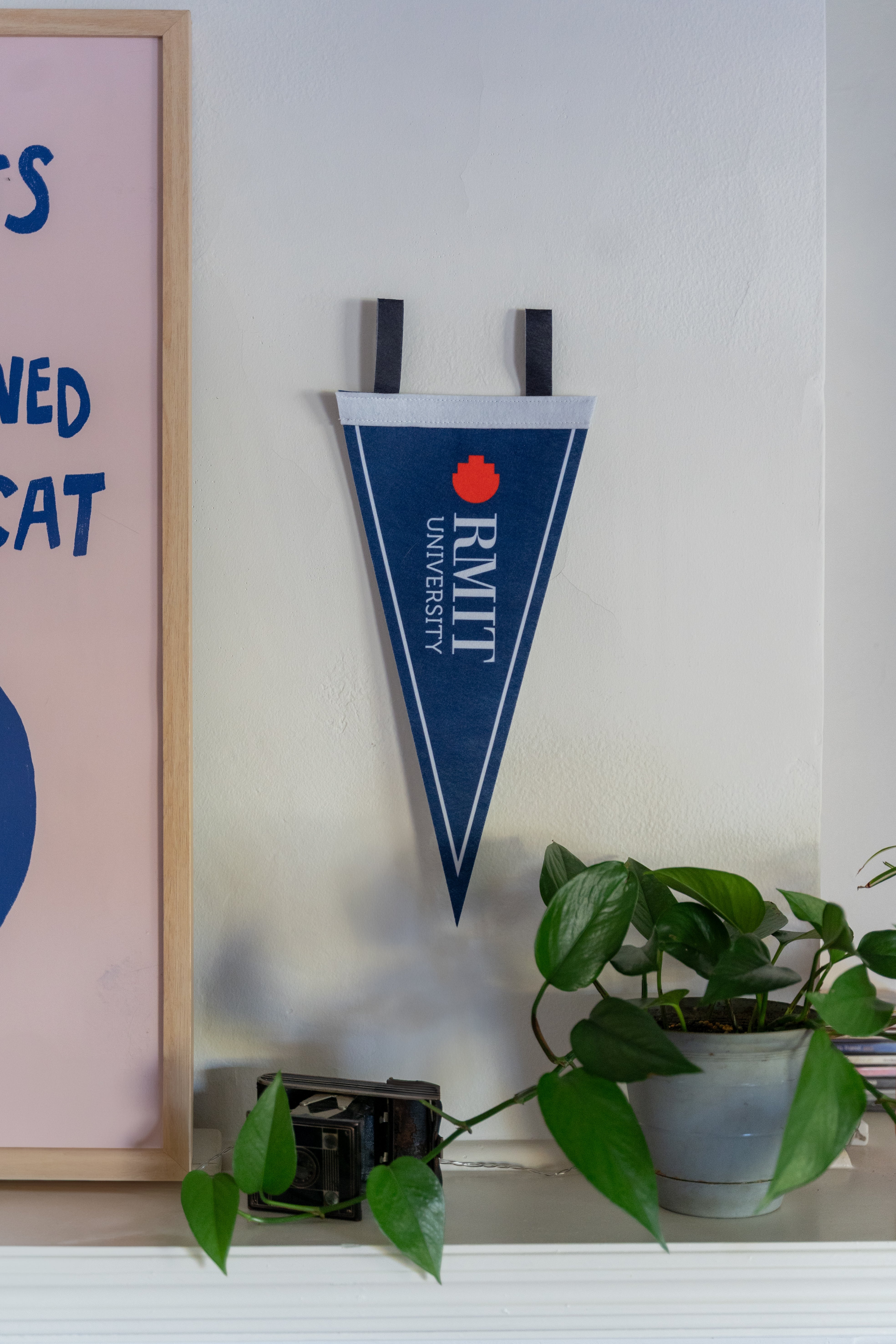 An RMIT Store felt pennant with a white border line hanging on a wall beside a framed artwork and a potted plant. The pennant features a blue background with a red pixel logo and white 'RMIT University' text, showcasing RMIT branding.