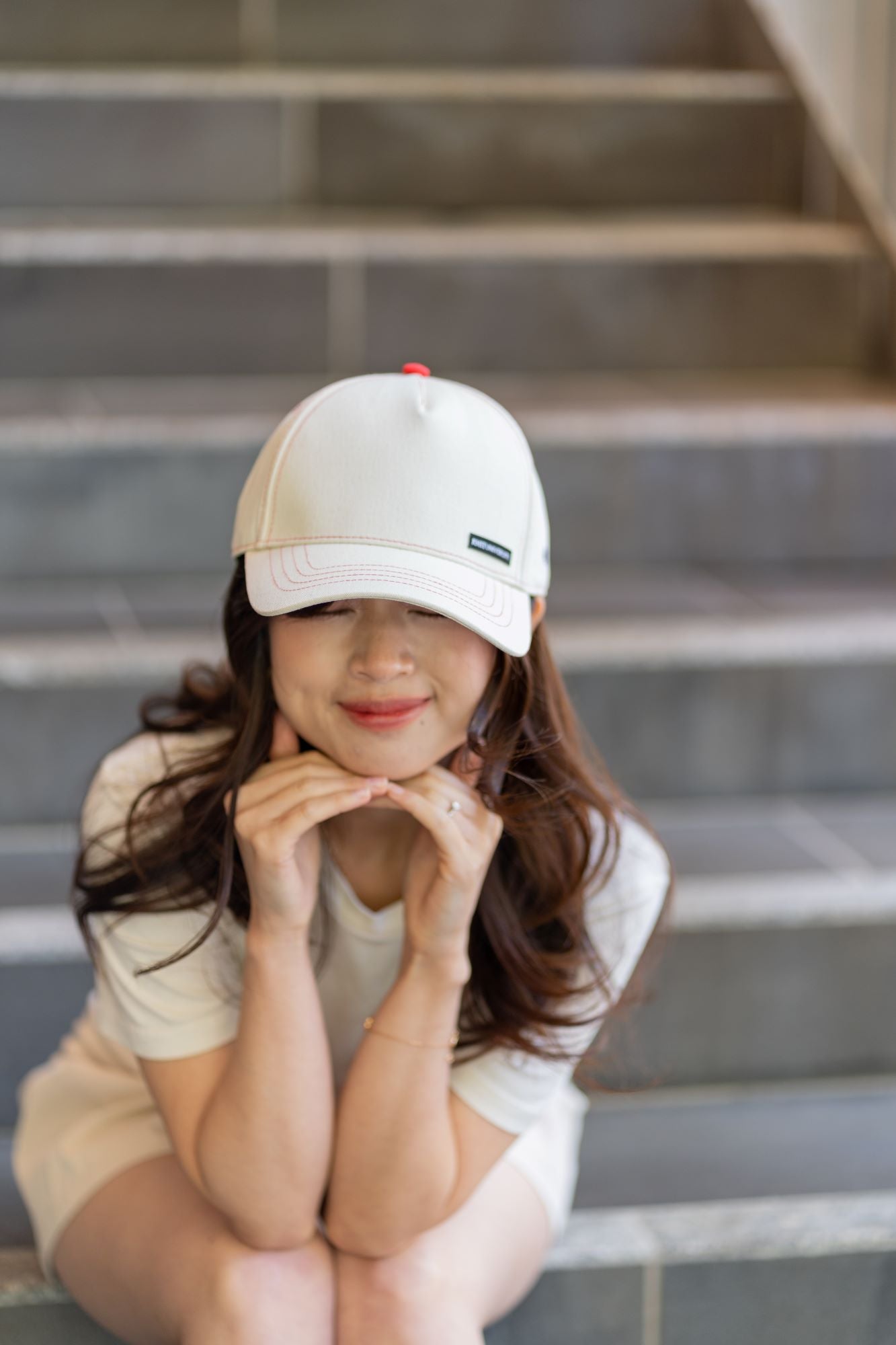 RMIT Store flex fit cap in light beige, adorned with red stitching and the RMIT pixel logo, worn by a model seated on stairs. The cap features a sleek design, ideal for casual wear.