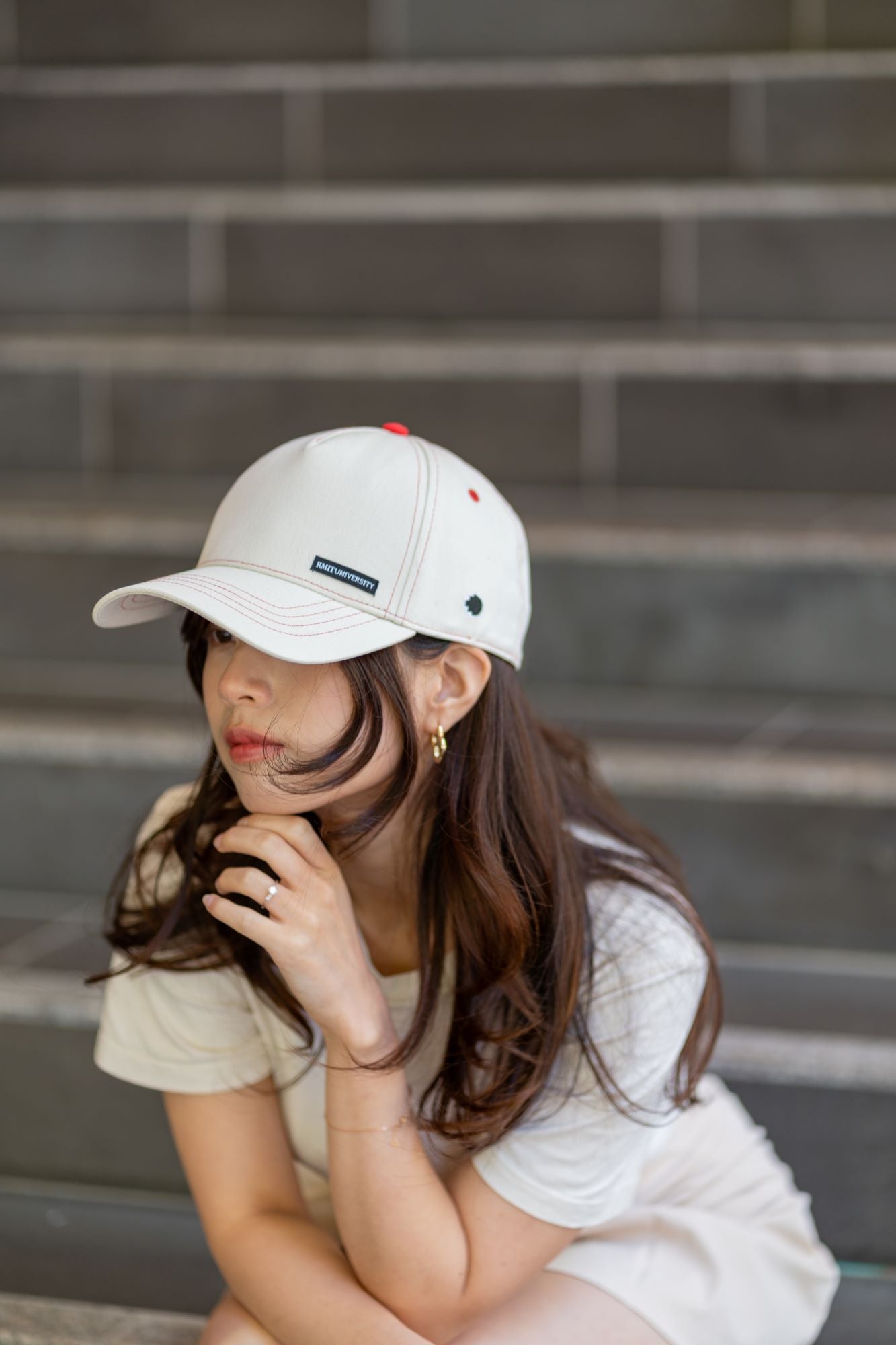 Light beige RMIT Store flex fit cap with red accents and pixel logo, styled by a model seated on stairs. The cap's minimalist design is complemented by earrings and relaxed attire.