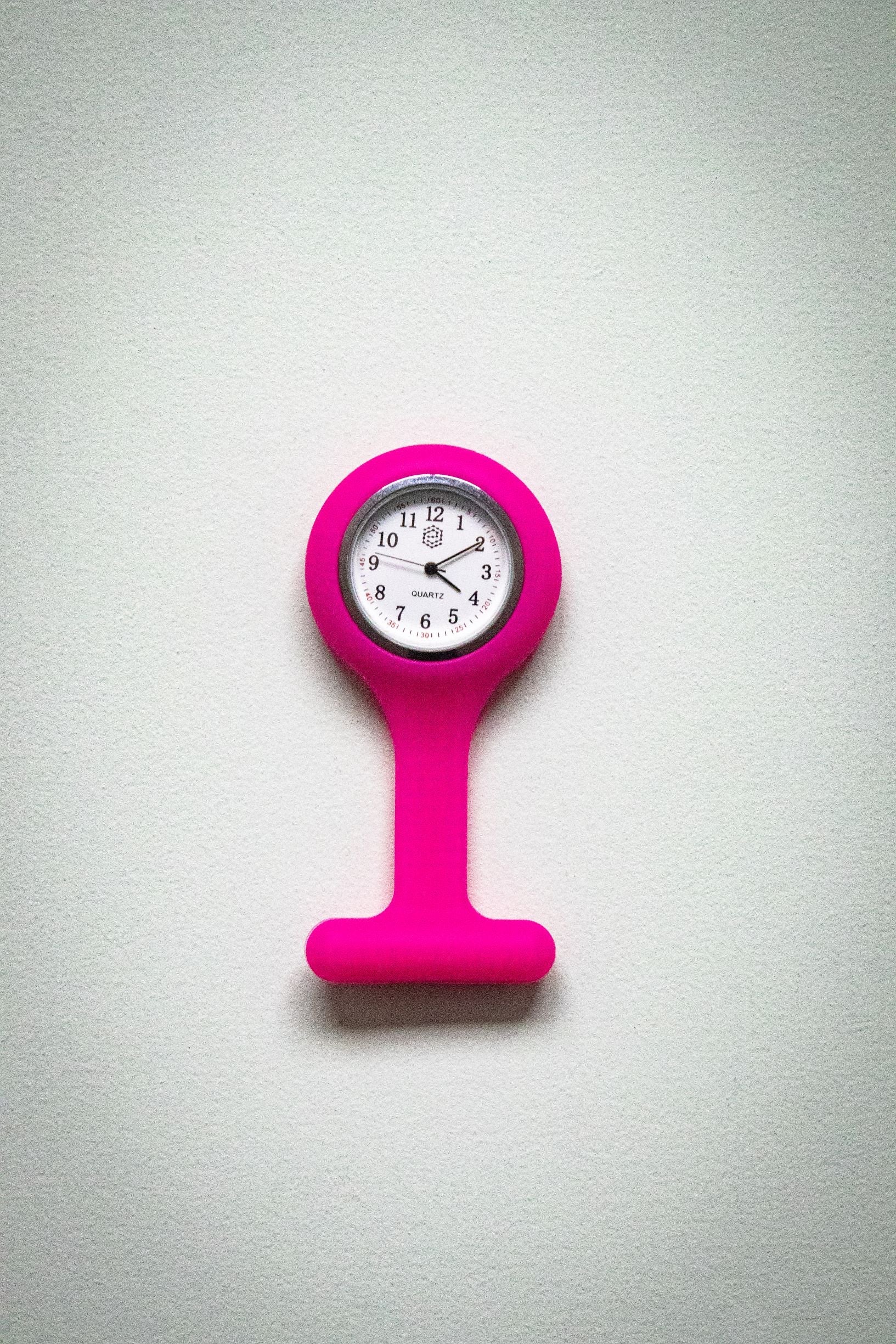 Upside-down front view of the RMIT Store magenta fob watch, displaying a white clock face with black numerals and three black hands inside a magenta silicone casing. 