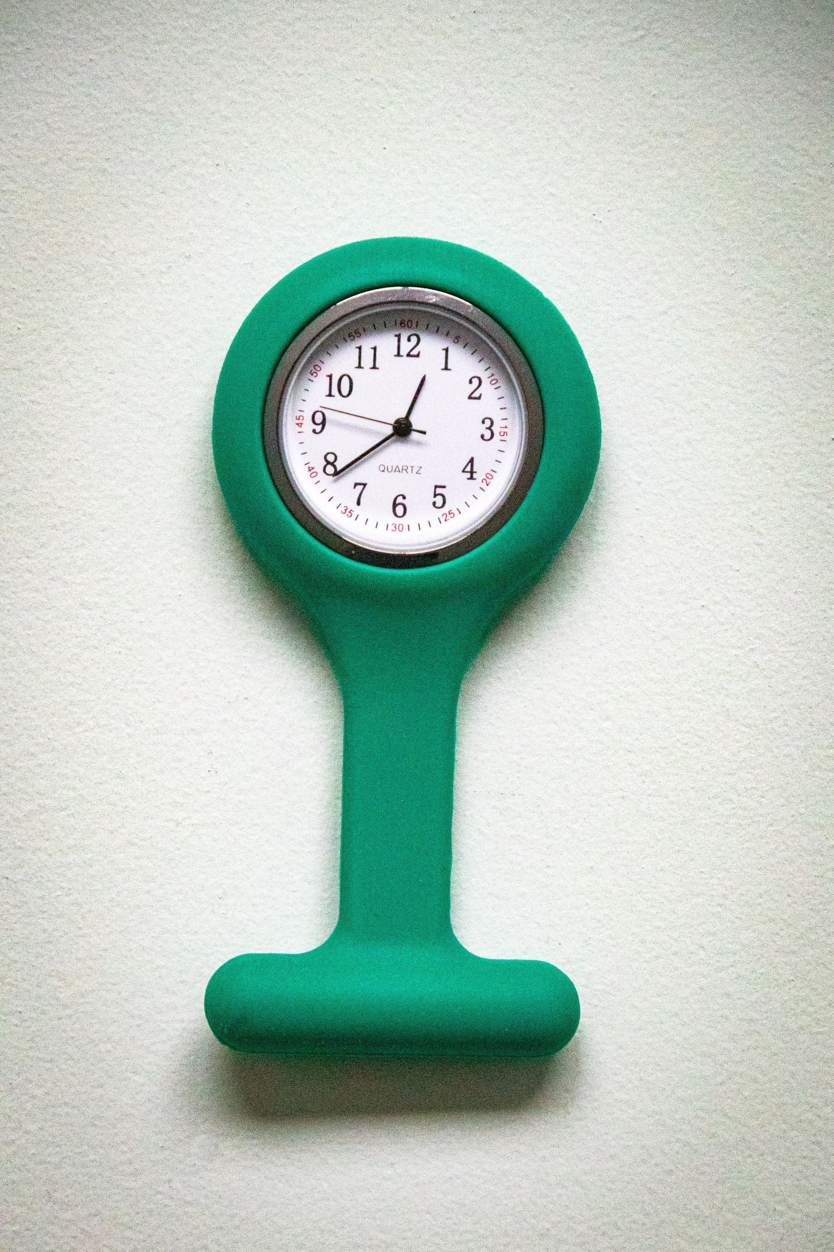 Upside-down front view of the RMIT Store teal fob watch, featuring a white clock face with black numerals and three black hands inside a teal silicone casing.