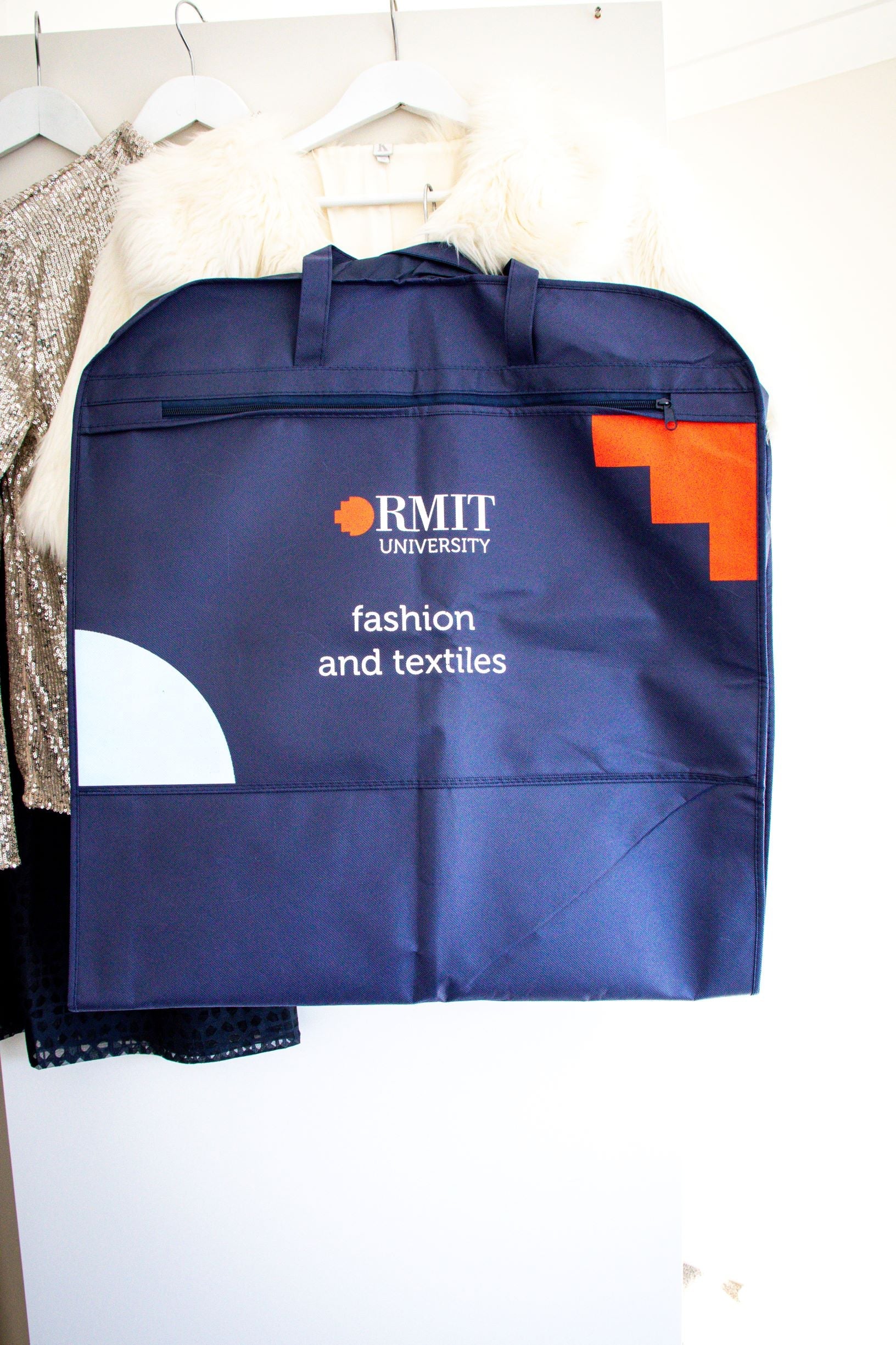 RMIT Garment Bag in navy, shown folded and hung among several garments on hangers, with the RMIT University logo in white and red prominently printed on the side, and the text “Fashion and Textile” written beneath the logo. Displayed against a plain white wall, this image highlights the compact fold and branded design of the product available from the RMIT Store.