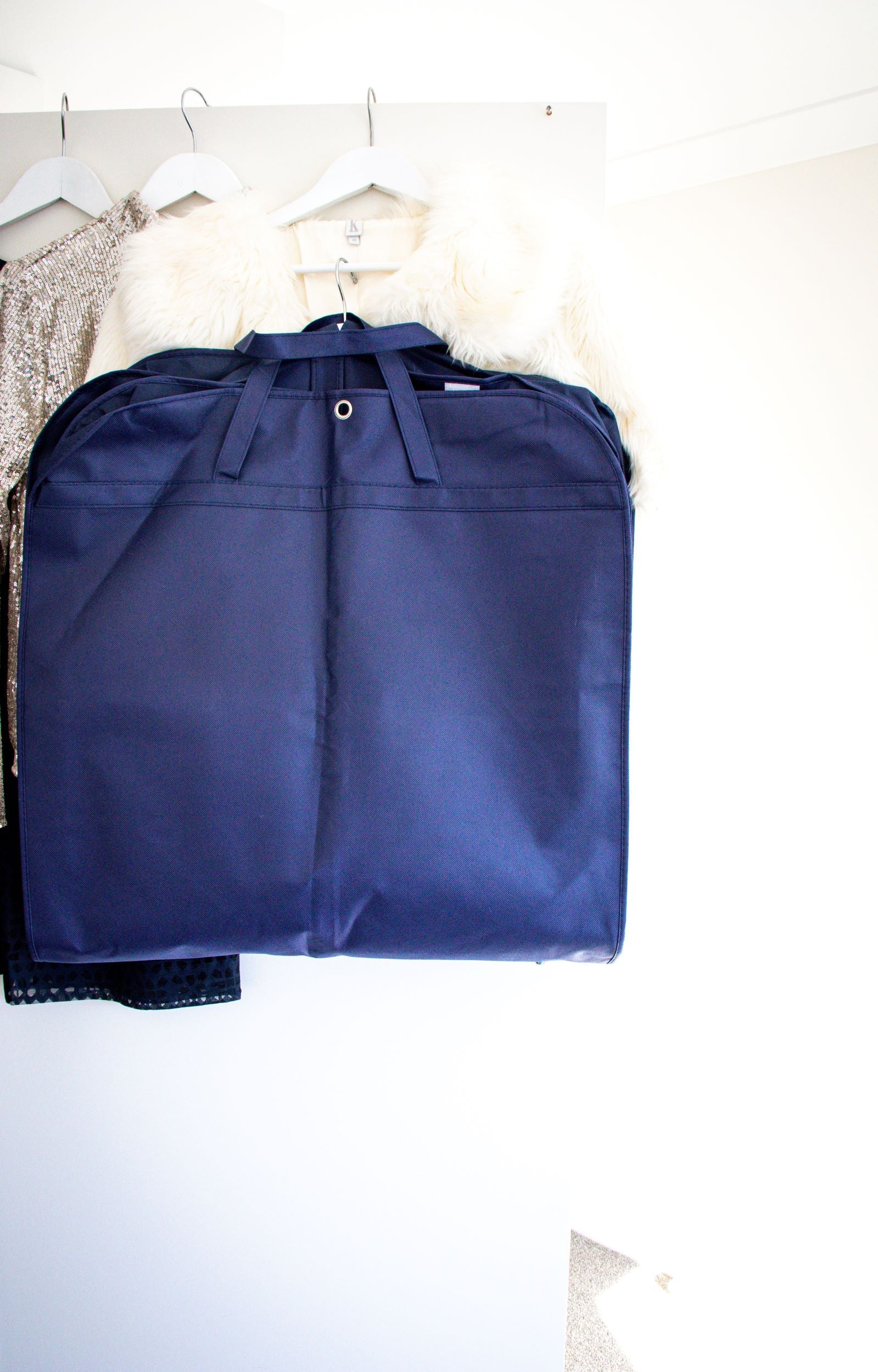 RMIT Garment Bag in navy, neatly folded and hung among several layered garments on hangers, with no visible branding or logo. The setting is a plain white wall background, and the image highlights the bag’s compact, durable design for protective garment storage. Available from the RMIT Store.