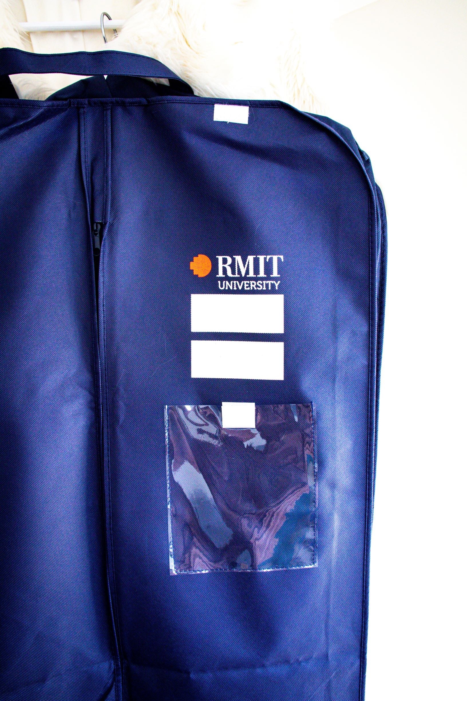 Front view of the RMIT Garment Bag in navy, shown hanging against a white wall. The “RMIT University” logo appears in white and red at the top right side of the bag. Below the logo are two blank white-labelled areas for writing, followed by a clear plastic pouch. A vertical zip runs down the centre of the bag. This official product is part of the RMIT Store collection.