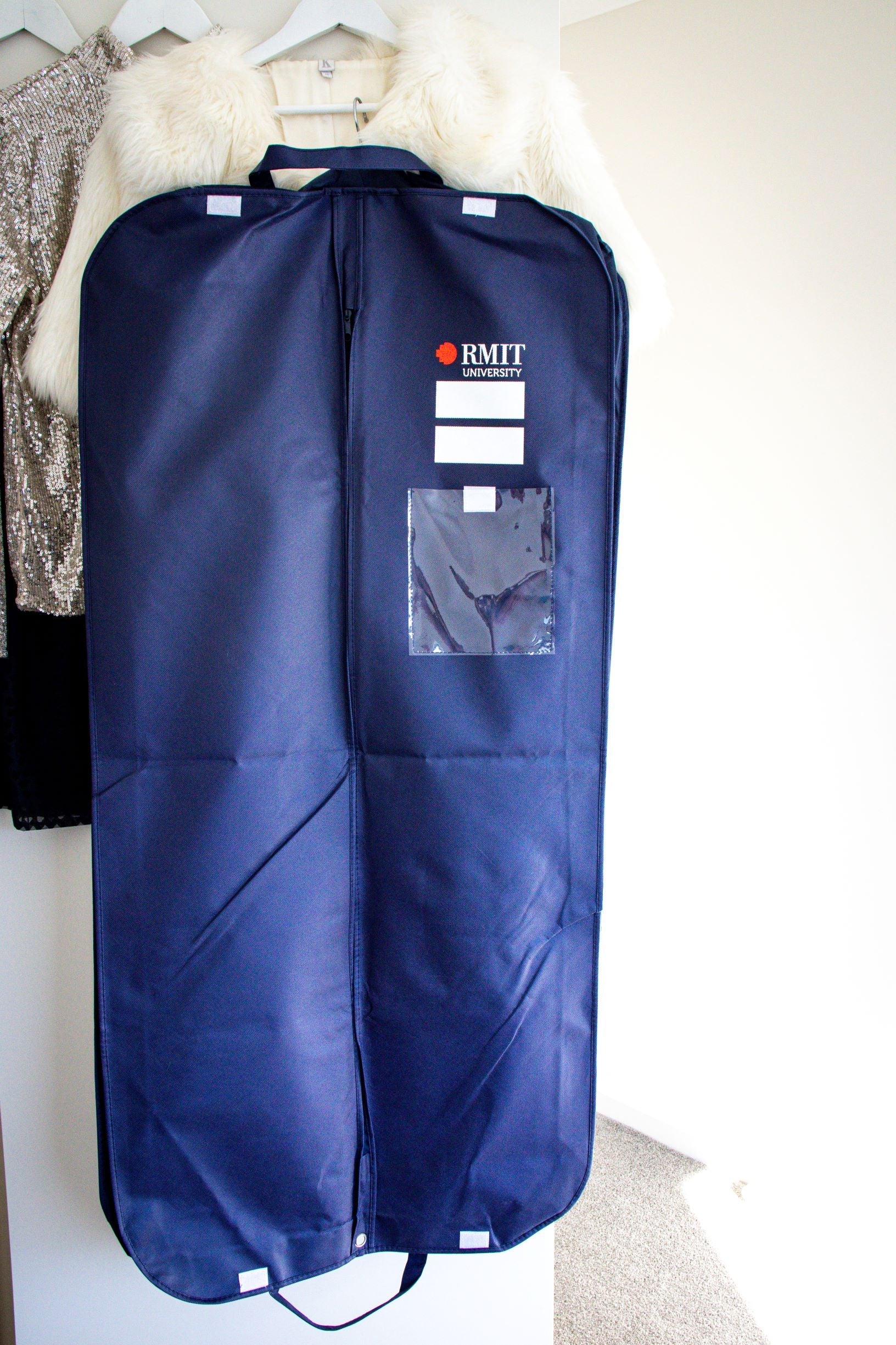 Full view of the RMIT Garment Bag in navy, displayed hanging against a white wall. The bag features the “RMIT University” logo in white and red on the top right side, with two blank areas beneath the logo for writing. A clear plastic pouch is located below the blank areas, and a central zip runs down the middle of the bag. The bag is shown in its entirety, highlighting its size and features as part of the RMIT Store collection.