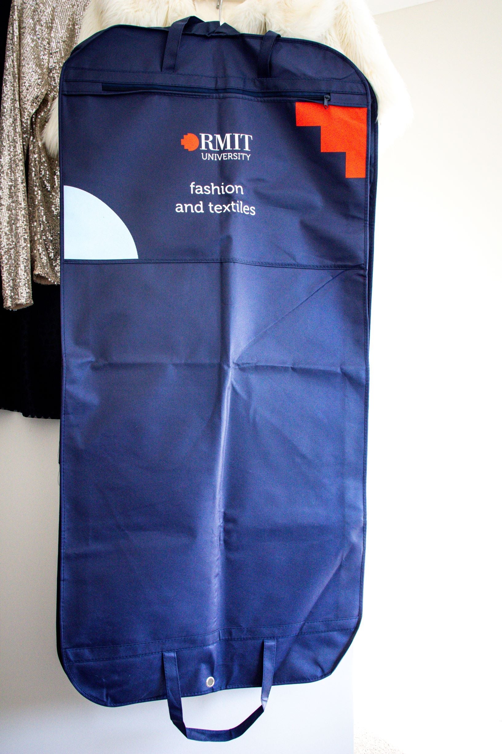 Close-up zoomed-in view of the rear of the RMIT Garment Bag in navy, focusing on the "RMIT University" logo in white and red at the top, with the words "Fashion and Textile" written underneath in white text. The image highlights the details of the branding and text on the bag's rear panel.