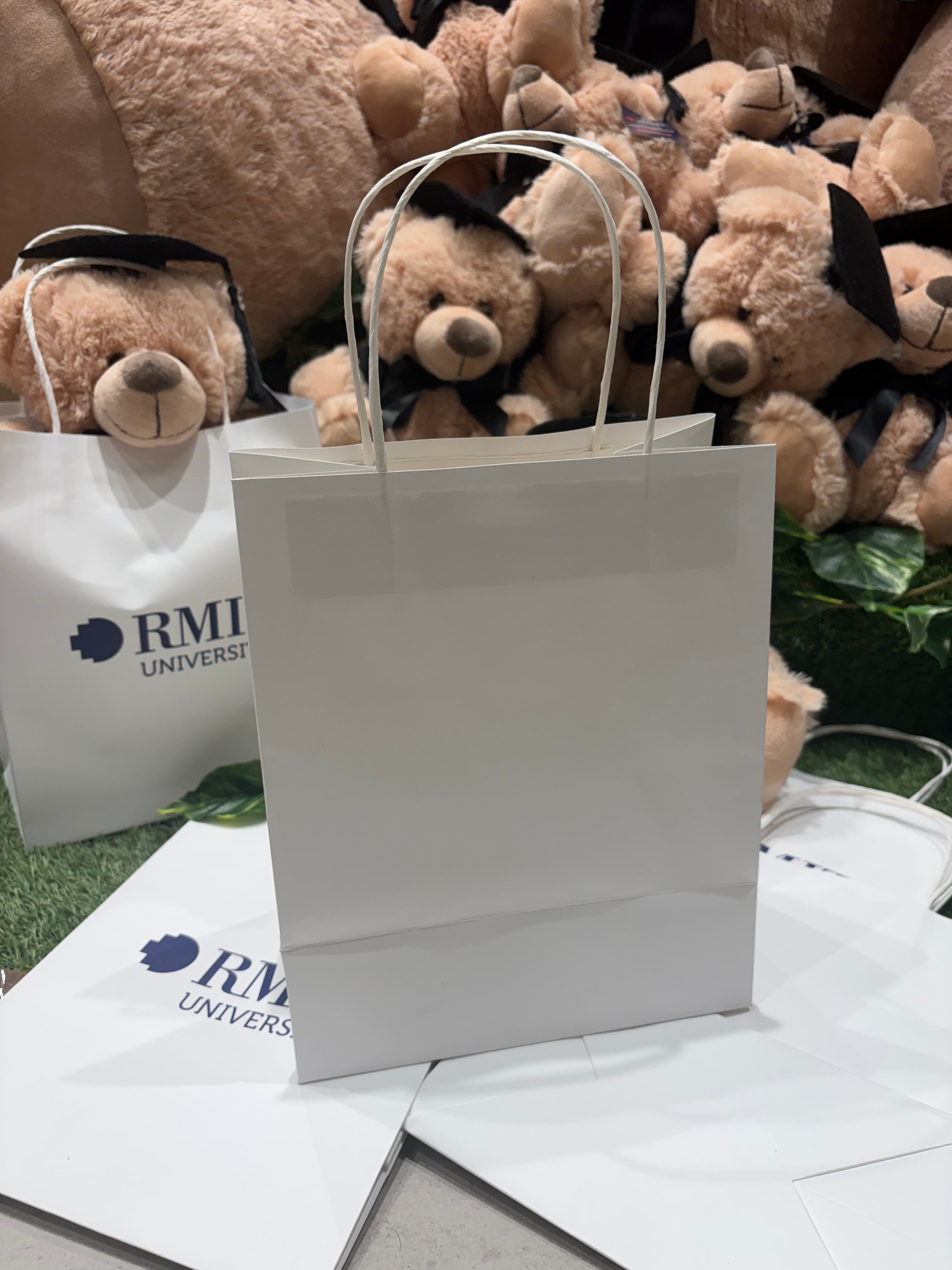 Rear view of a plain white gift bag with twisted handles displayed on a stack of folded RMIT University gift bags, with graduation teddy bears positioned in the background inside the RMIT Store.